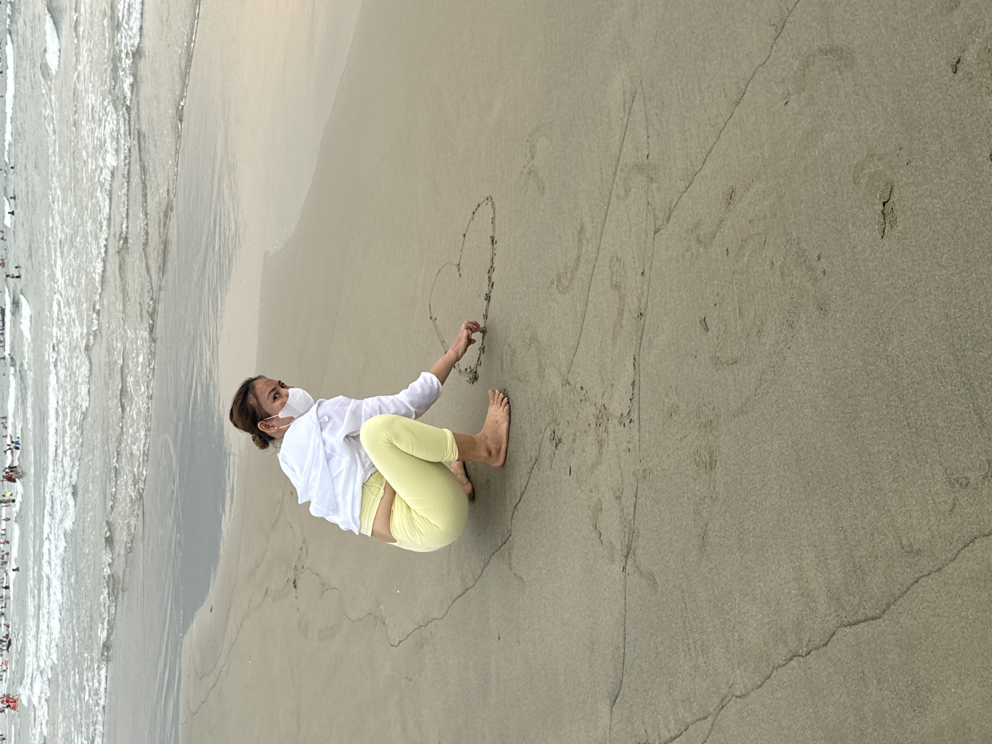 A woman wearing a face mask, white shirt, and light yellow leggings is crouched on a sandy beach, drawing a heart in the sand. The ocean is visible in the background, with people near the water's edge. The location is likely a beach in Đà Nẵng, Vietnam.