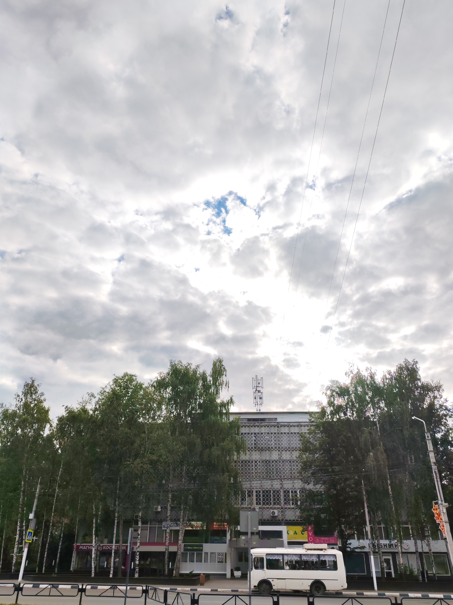 A white bus is parked on the side of a street in Chaikovsky, Russia, with a building and trees in the background. The bus appears to be occupied by passengers. The building behind the bus is a multi-story structure with a patterned facade, and there are trees lining the street in front of it. The sky is overcast with some blue patches visible. Power lines are visible across the top of the image.