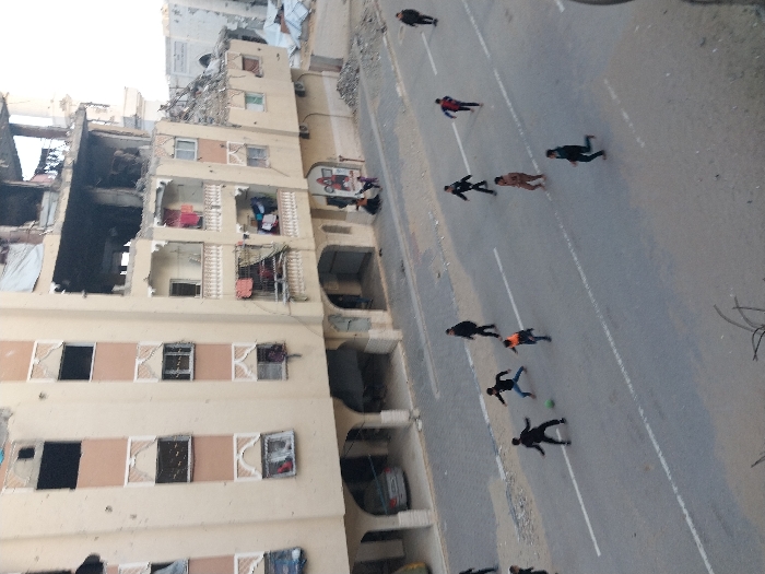 An elevated view depicts a street scene in Khan Yunis, Palestinian Territory. The foreground shows an asphalt road with faded white dashed lane markings. Approximately nine young males are actively playing soccer in the street, with one light green ball. They are dressed in casual dark attire, with figures in various stages of running and kicking.
To the left of the road, a large, multi-story residential building dominates the background. This light-beige structure exhibits widespread damage, including numerous bullet holes on its facade, broken and missing windowpanes, and exposed interior sections on its upper and right sides, indicative of structural collapse. Despite the damage, laundry hangs on several balconies, suggesting partial habitation or recent presence. A silver sedan is parked beneath an arched ground-floor entrance.
Further to the right, a second, heavily damaged building is visible, with significant debris piles at its base and on the adjacent ground. A white architectural feature, possibly part of a mosque minaret or dome, is visible behind this building. A small group of 3-4 individuals, including adults and children, stands near the entrance of the main building, observing the scene. One additional male figure walks along the far right edge of the road, moving away from the central activity. The scene is illuminated by natural daylight, indicating daytime.