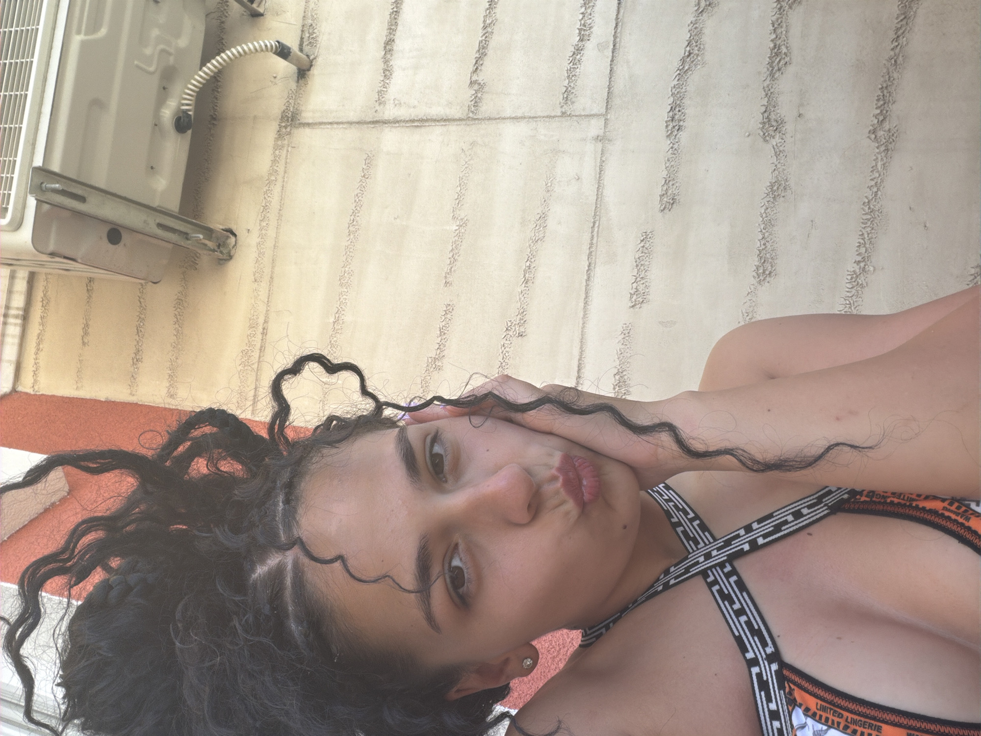 A young woman with dark, curly hair and a diamond stud earring is pictured. She is wearing a bikini with a black and white geometric pattern and orange trim, which has "LIMITED LINGERIE" printed on it. Her right hand is touching her face, and her lips are pursed. She is lying down outdoors, with an air conditioning unit mounted on a textured wall behind her. The wall appears to be made of concrete blocks with vertical, decorative grooves. The lighting suggests it is daytime, possibly sunny, given the shadows and bright illumination. The overall impression is one of leisure, perhaps in a warm climate. The background hints at a location in Nesebar, Bulgaria.