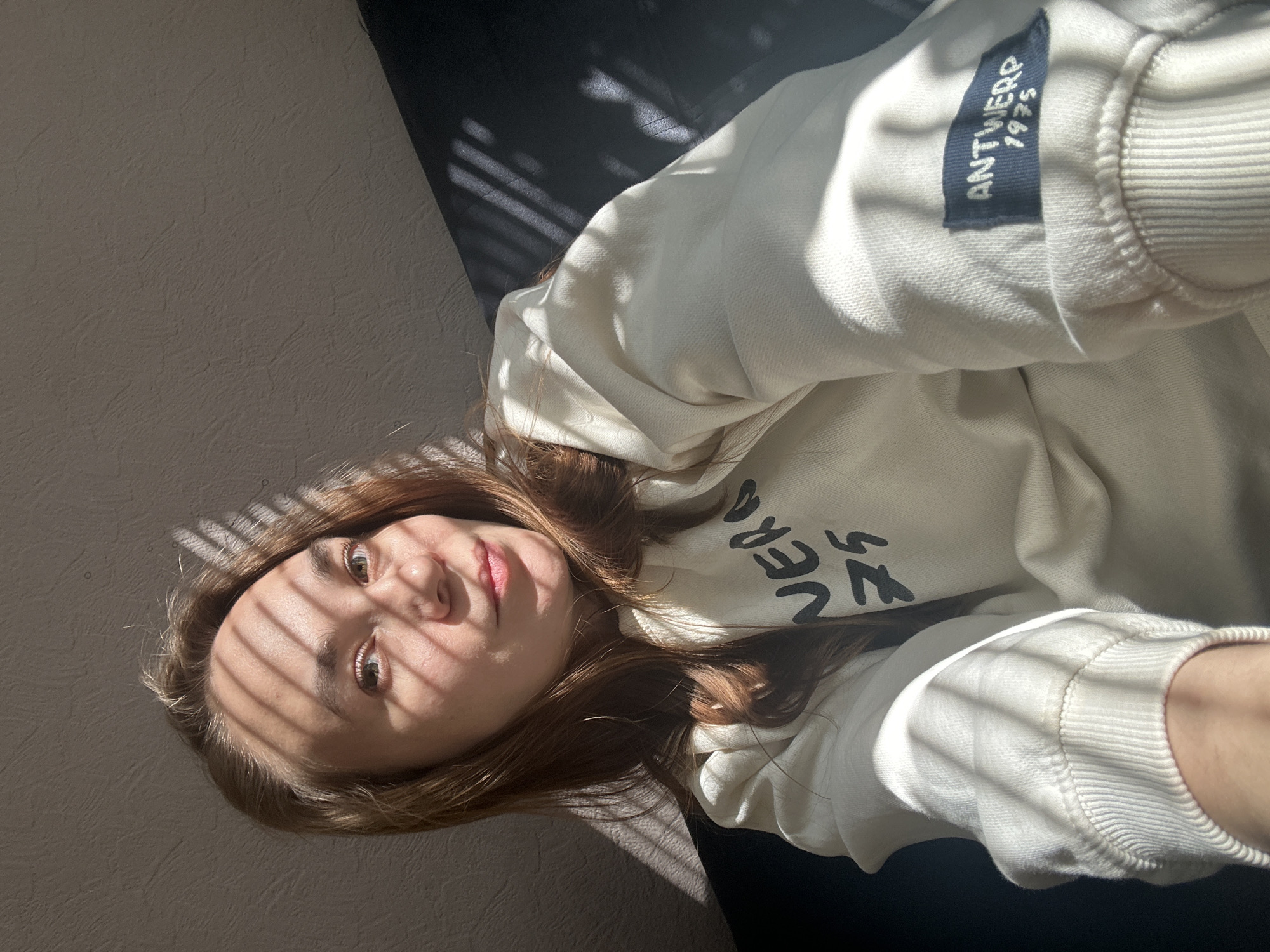 A young woman is lying down indoors, possibly in a room, with sunlight filtering through blinds creating shadows across her face. She is wearing a light-colored sweatshirt with the word "ANTWERP 1975" on the sleeve and an unidentifiable word across the front. The background is textured and appears to be a wall and a dark surface. There is no information given in the image that suggests the location is Chaikovsky, Russia.