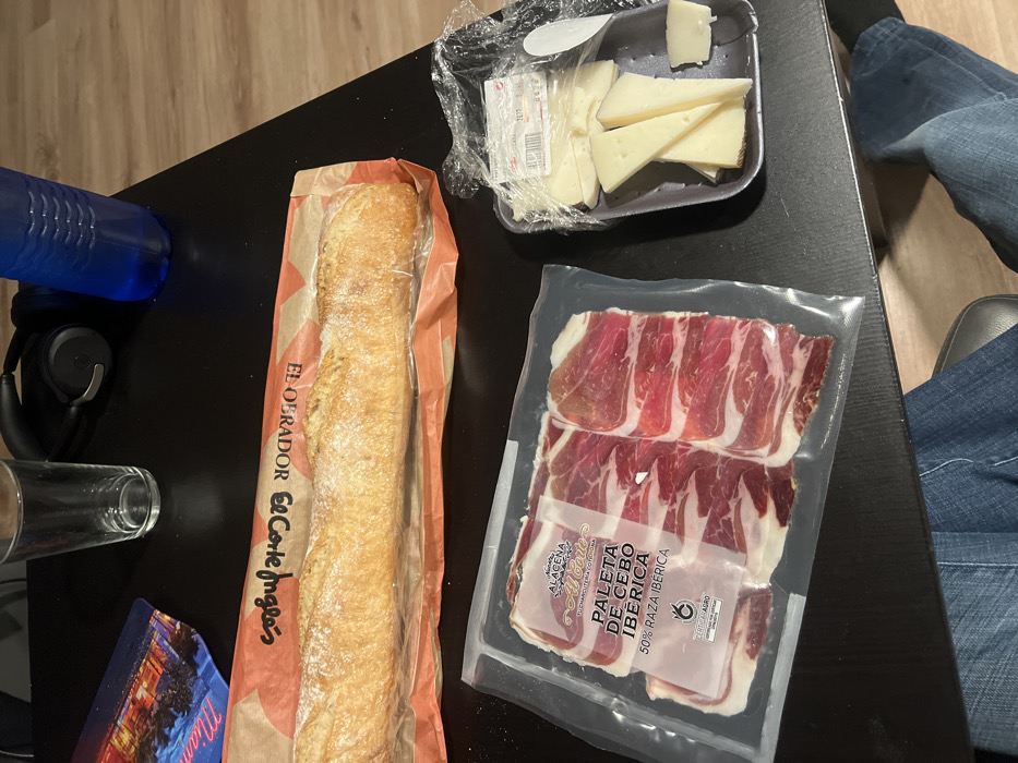 A baguette in its branded paper bag, cheese slices in a plastic container, and packaged ham slices, specifically "Paleta de Cebo Ibérica 50% Raza Ibérica," rest on a dark surface. Accompanying these food items are a blue water bottle, headphones, a glass, and a souvenir card featuring a skyline. The setting appears casual, possibly a table where a meal or snack is being prepared.
