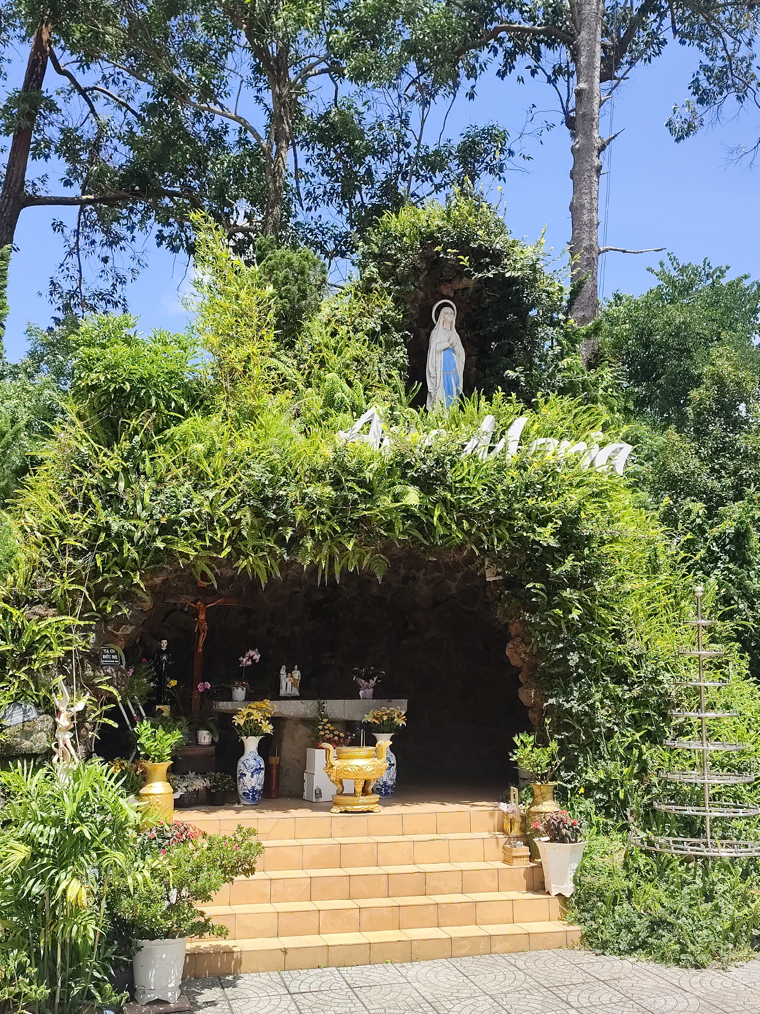 The image shows a grotto dedicated to Mary, located at the Dinh 2 (Palace II) in Da Lat, Vietnam. The grotto is built into a hillside and covered with lush greenery, with a statue of Mary at the top. Below the statue, a carved inscription reads "AN MA" in white lettering, partially obscured by foliage. Steps lead up to the entrance of the grotto, where an altar is set up. On the altar, religious figurines and offerings are visible, including a crucifix, statues of saints, vases of flowers, and a golden incense burner. Various plants and potted flowers surround the grotto and line the steps, contributing to the natural and serene atmosphere. The sky above is a clear blue, and sunlight illuminates the scene, casting shadows on the ground.