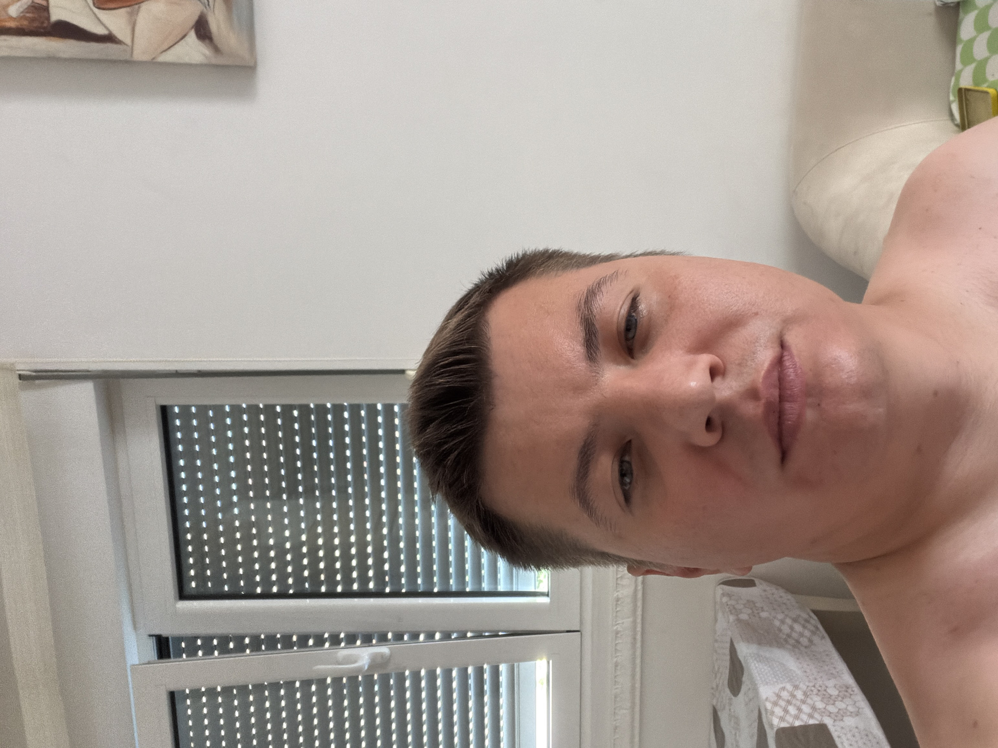 This is a selfie of a person lying down, with their head tilted back. The person has fair skin and short brown hair. They are looking directly at the camera with a neutral expression. The background is out of focus but appears to be an indoor setting with a white wall and a window with white blinds. There is also a hint of a painting on the wall in the top left corner, and a light-colored couch or chair in the background on the right. The lighting suggests it could be daytime. The image is oriented sideways, suggesting it was taken with the camera held vertically. The setting is likely a home or hotel room in Budva, Montenegro, given the location context. No text is visible in the image.