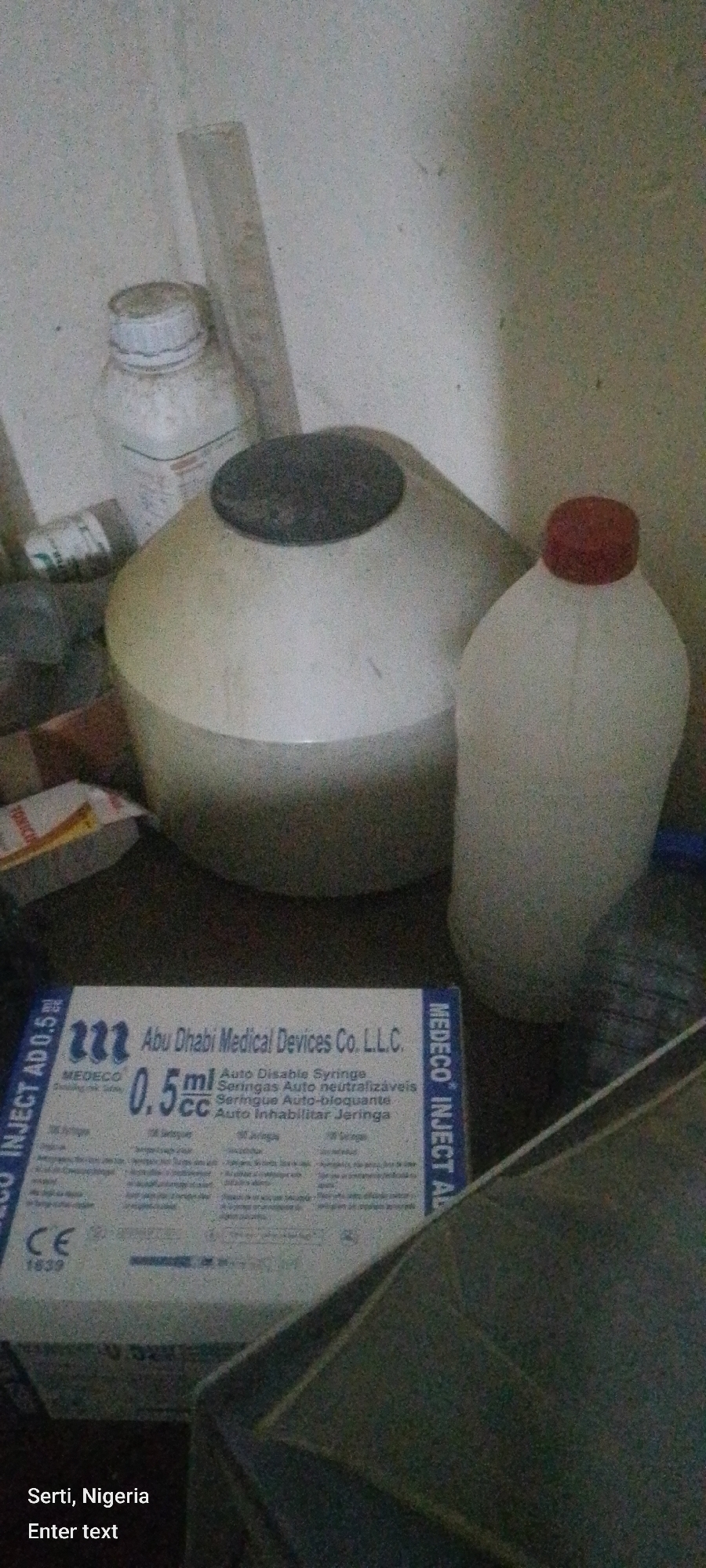 The image depicts a cluttered, indoor scene with various medical supplies and containers. In the foreground, a box of "MEDECO INJECT AD 0.5 ml CC Auto Disable Syringe" manufactured by Abu Dhabi Medical Devices Co. L.L.C. is prominently displayed. To the right of the box is a translucent plastic bottle with a red cap, and to the left, a large, off-white cylindrical container with a dark lid. Behind these items, a white bottle with a white cap is visible, along with other indistinct objects and what appears to be a ruler or measuring stick leaning against the wall. The background consists of a textured, light-colored wall. The lighting is dim, casting shadows and contributing to the overall grainy quality of the image. Text at the bottom of the image indicates the location as "Serti, Nigeria".