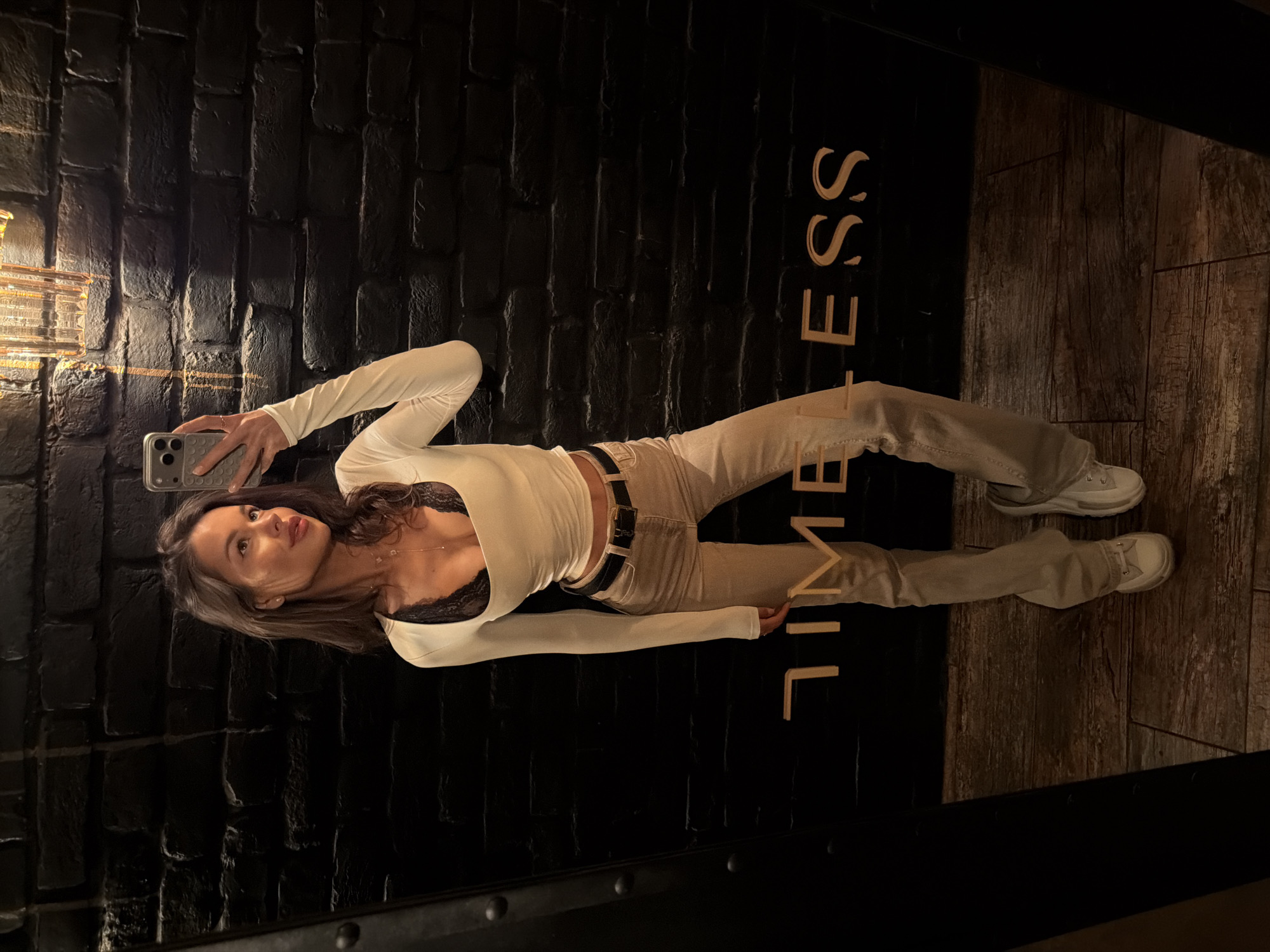 An indoor mirror selfie captures a young woman posing in a stylish setting. She holds a smartphone with a textured grey case in her left hand, taking the picture. She is dressed in a fitted, off-white long-sleeved top, revealing a black lace bra beneath, light beige trousers, a black belt, and white sneakers. Her dark brown hair cascades over her shoulders, and she looks towards the camera with a neutral, confident expression.
The background behind her features a dark, brick-patterned wall on the left, adorned with a gold accent strip and a decorative light fixture. To her right, the surface transitions to dark wooden paneling, likely a floor reflected in the mirror. Partially visible in gold lettering on the dark wall is the word "TIMELESS," oriented vertically. The lighting is dim, suggesting an evening or indoor artificial light setting.
Location context: Moscow, Russia.