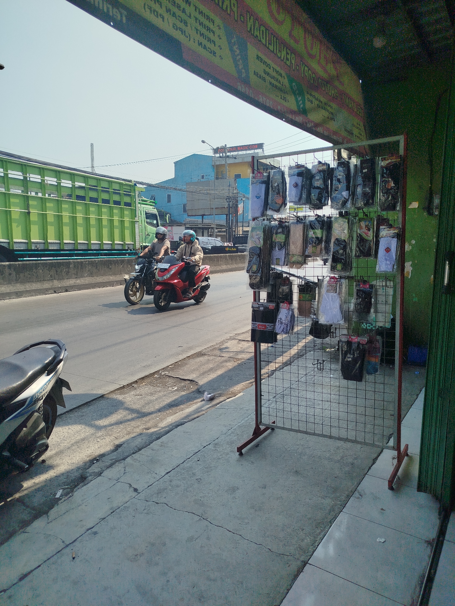 The image shows a street scene in Mranggen, Indonesia, with a focus on a roadside stall selling various items, likely accessories. The stall displays numerous packages of what appear to be socks or similar textile goods hanging on a wire rack. In the background, a large green truck is driving on a road, and two individuals are riding on motorcycles on the same road. The environment appears to be urban, with buildings visible in the distance and a sidewalk in the foreground. The lighting suggests it is daytime, possibly morning or afternoon, with shadows cast by the stall and objects on the sidewalk.