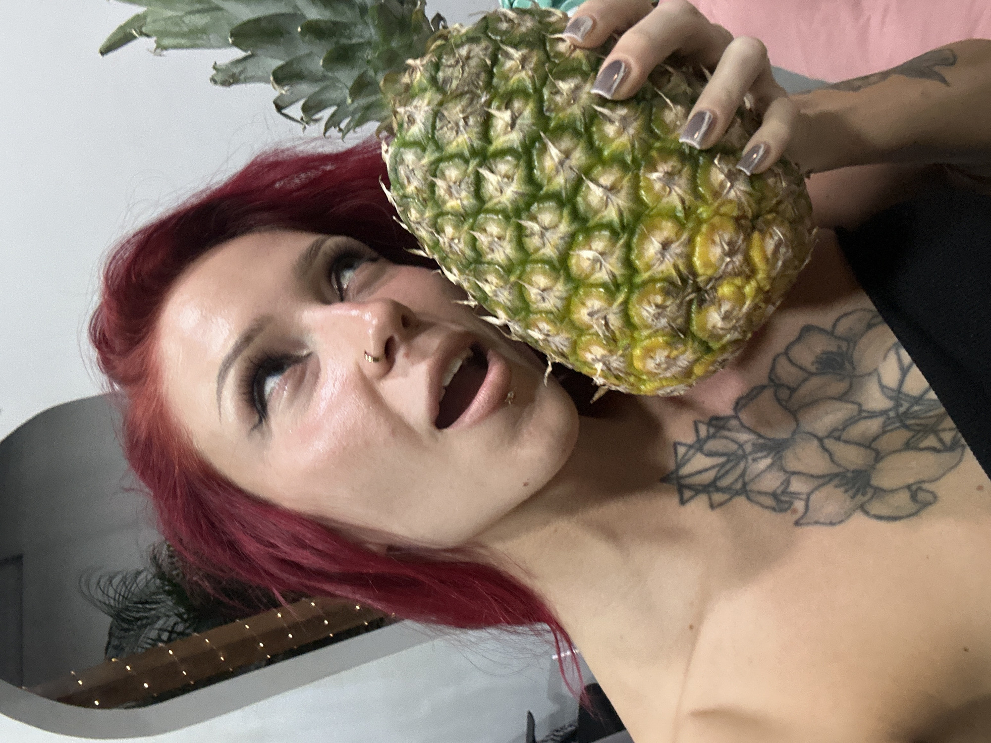 An angled, close-up portrait features a woman with vibrant red hair holding a large pineapple to the side of her face. She has prominent eye makeup, including dark eyeliner and mascara, and a small ring piercing in her left nostril. Her mouth is slightly open, and her eyes are looking upwards, conveying an expressive and somewhat playful or surprised look. A detailed floral tattoo is visible on her right shoulder/upper chest, and another tattoo is partially seen on her left arm. Her fingernails are painted with a light grey or mauve polish accented with white tips. The pineapple itself is green and yellow, held firmly by her right hand. The background appears to be an indoor setting with a plain light-colored wall or ceiling, a hint of a soft pink surface, and some blurry ambient lights. The overall lighting suggests artificial illumination. This image was captured in North Kuta, Indonesia.