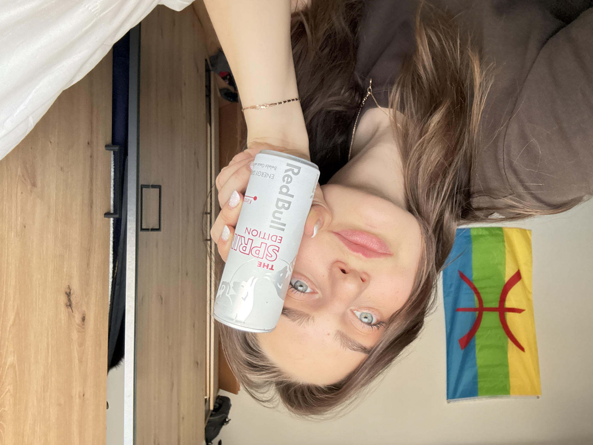 An indoor, waist-up shot shows a young woman holding a Red Bull can and looking directly at the camera. She has long, wavy brown hair with lighter highlights framing her face, striking blue-green eyes, and a neutral expression with slightly parted lips. She wears a dark, possibly brown, long-sleeved top and a delicate chain bracelet on her right wrist, along with a thin necklace. Her nails are manicured with white tips.
She holds a silver Red Bull can with both hands. Text visible on the can includes "Red Bull," "THE SPRING EDITION," and "KIRSCHE" (Cherry), along with the German slogan "BELEBT GEIST UND KÖRPER" (Invigorates mind and body).
In the background, to the right of her head, a Berber flag is displayed, featuring vertical stripes of blue, green, and yellow, with a prominent red "ⵣ" (Yaz) symbol in the center. To her left, wooden panels, likely part of a wardrobe or door, are visible. A section of white fabric, possibly a curtain or clothing, can be seen in the upper left corner. The lighting suggests it is daytime. While the image is provided with the location context of Hainburg an der Donau, Austria, no specific visual cues within the image itself confirm this location.