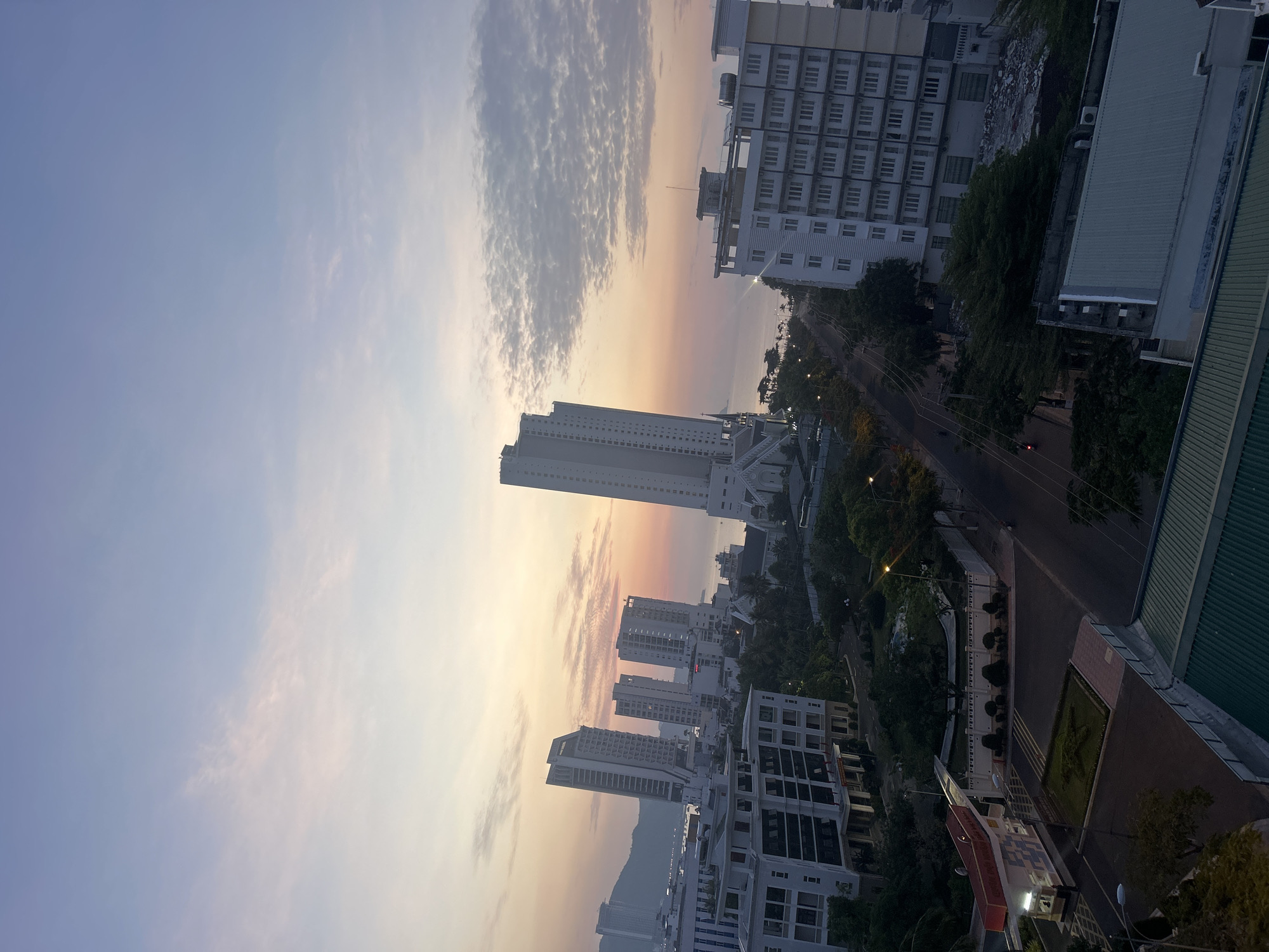 Captured from an elevated perspective, the image portrays a cityscape in Bac Nha Trang Ward, Vietnam, at either sunrise or sunset. The sky displays a gradient of colors from light blue to orange, with scattered clouds. Several tall buildings dominate the skyline, including one exceptionally high-rise structure. The streets below are lined with trees and streetlights, showing minimal vehicular traffic. To the right, the edges of buildings and green foliage are visible, potentially indicating a park or garden area. The scene is devoid of any observable human activity.