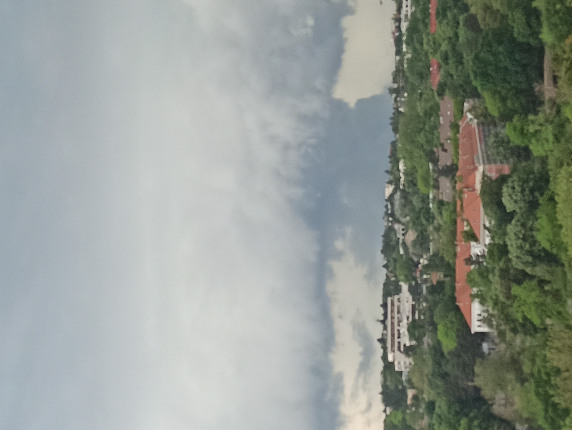 The image shows a view from what appears to be a high vantage point, likely in Vozdovac Urban Municipality, Serbia. The sky is overcast with grey clouds dominating the left side of the image. On the right side, there is dense vegetation with trees and bushes in varying shades of green. Interspersed within the greenery are buildings, some with red tile roofs and others appearing to be white apartment buildings. The architectural style and the density of buildings suggest an urban or suburban area nestled among the trees. There are no people or evident interactions visible in the scene. The general impression is of a hillside or elevated area overlooking a residential area with abundant foliage.