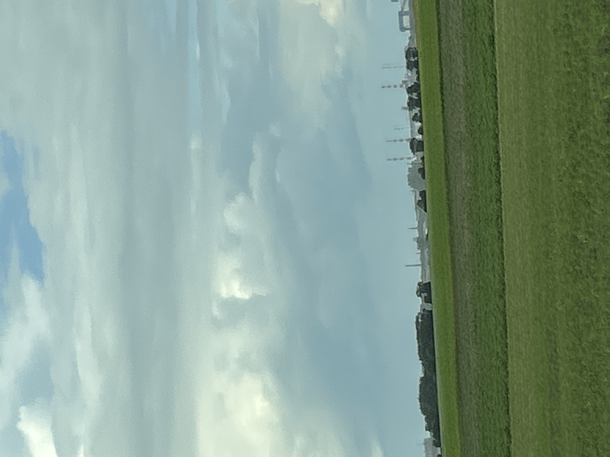 The image depicts a wide, horizontal landscape with a focus on agricultural fields and an industrial background under a cloudy sky. In the foreground, there are two distinct bands of green grass. The lower band is a vibrant, lighter green, while the band above it is a darker, more muted green. These fields appear to be well-maintained, suggesting agricultural use.
In the middle ground, a slightly elevated line of dark green vegetation runs horizontally across the frame, potentially a hedgerow or a line of trees. Behind this vegetation, a distant industrial area is visible. This industrial zone features multiple tall, slender chimneys or smokestacks, some with visible markings or bands. There are also several low-rise industrial buildings and structures, including what appears to be a large, grey crane or gantry structure on the far right. The overall impression of the industrial area is one of significant scale and activity.
The sky above is filled with a dense layer of grey and white clouds, with patches of blue sky visible in the upper portion of the image. The lighting suggests an overcast day, possibly with some sunlight filtering through the clouds, creating a diffused, soft light.
The specific location in Красненскі сельскі Савет, Belarus, is characterized by this blend of agricultural land and industrial development, a common feature in many rural or semi-urbanized areas where industry coexists with farming. The scene is static and devoid of any visible human presence or activity.