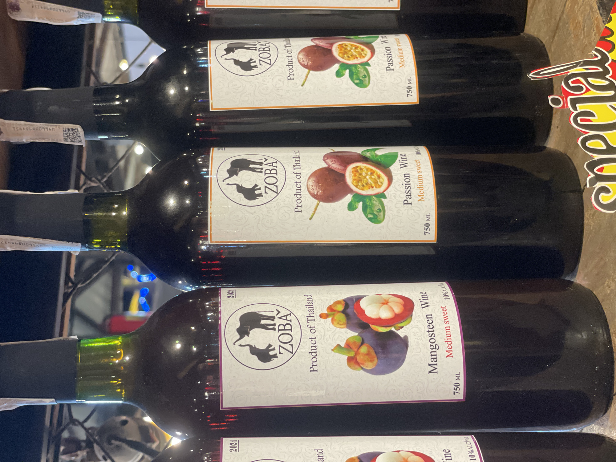 The image shows a close-up of several dark glass wine bottles lined up, likely in a retail setting in Pattaya City, Thailand. The bottles have white labels featuring a silhouette of two elephants, the brand name "ZOBA," and the text "Product of Thailand." Some labels depict images of mangosteens and are labeled "Mangosteen Wine," while others show passion fruits and are labeled "Passion Wine." All wines appear to be 750 ML with 10% ALC. In the foreground, a brightly colored "Special" sign is partially visible. The background is blurred, showing hints of lights and structures, suggesting an indoor or covered outdoor market or shop.