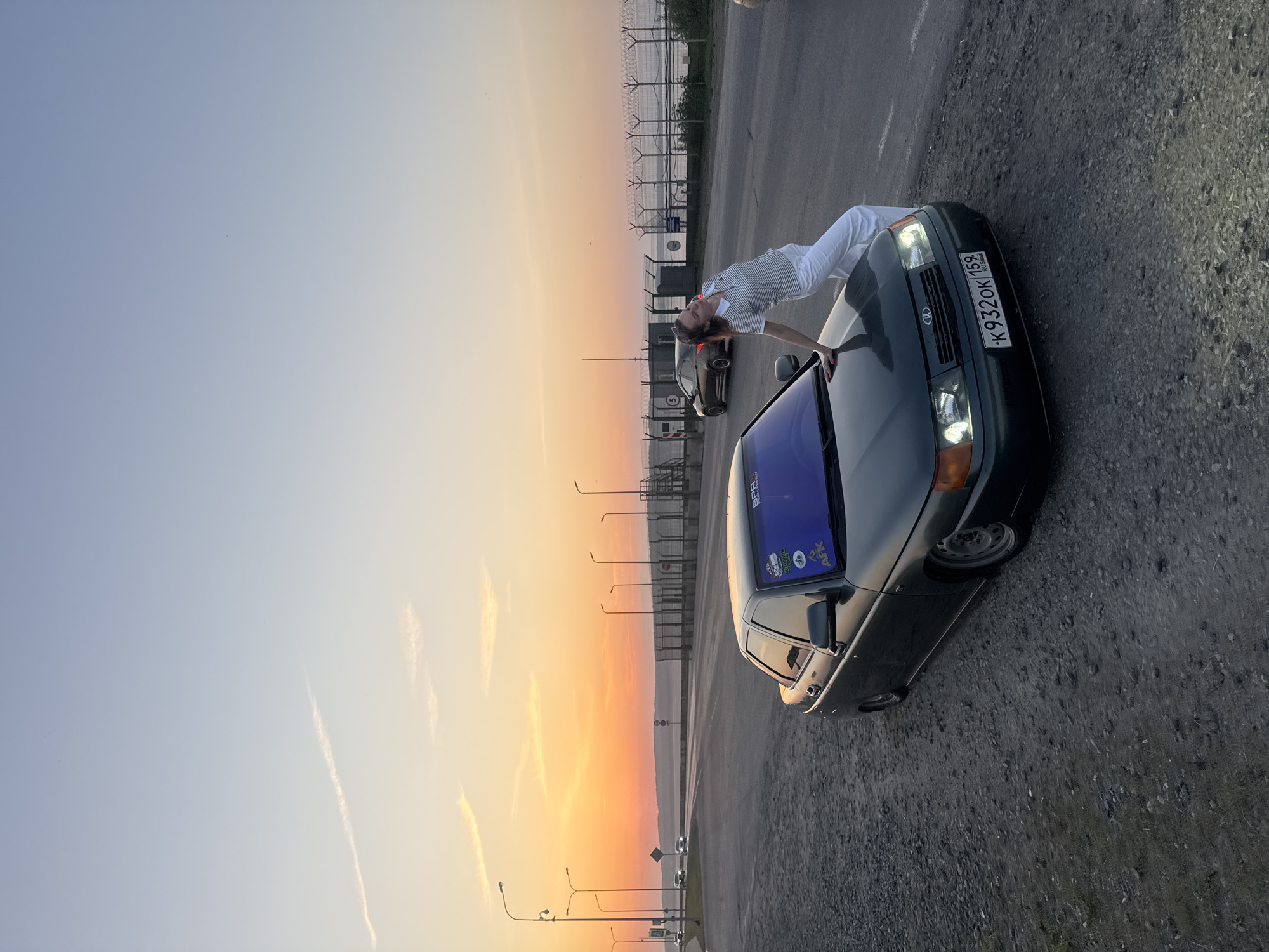 A photograph shows a person in white clothing leaning over a dark-colored car. The license plate number "K932OK 159" is visible on the front of the car. In the background, there is a sunset sky with a gradient of orange and yellow. There are streetlights and a security fence in the distance. The location is consistent with Chaikovsky, Russia, as indicated by the license plate region code "159".
