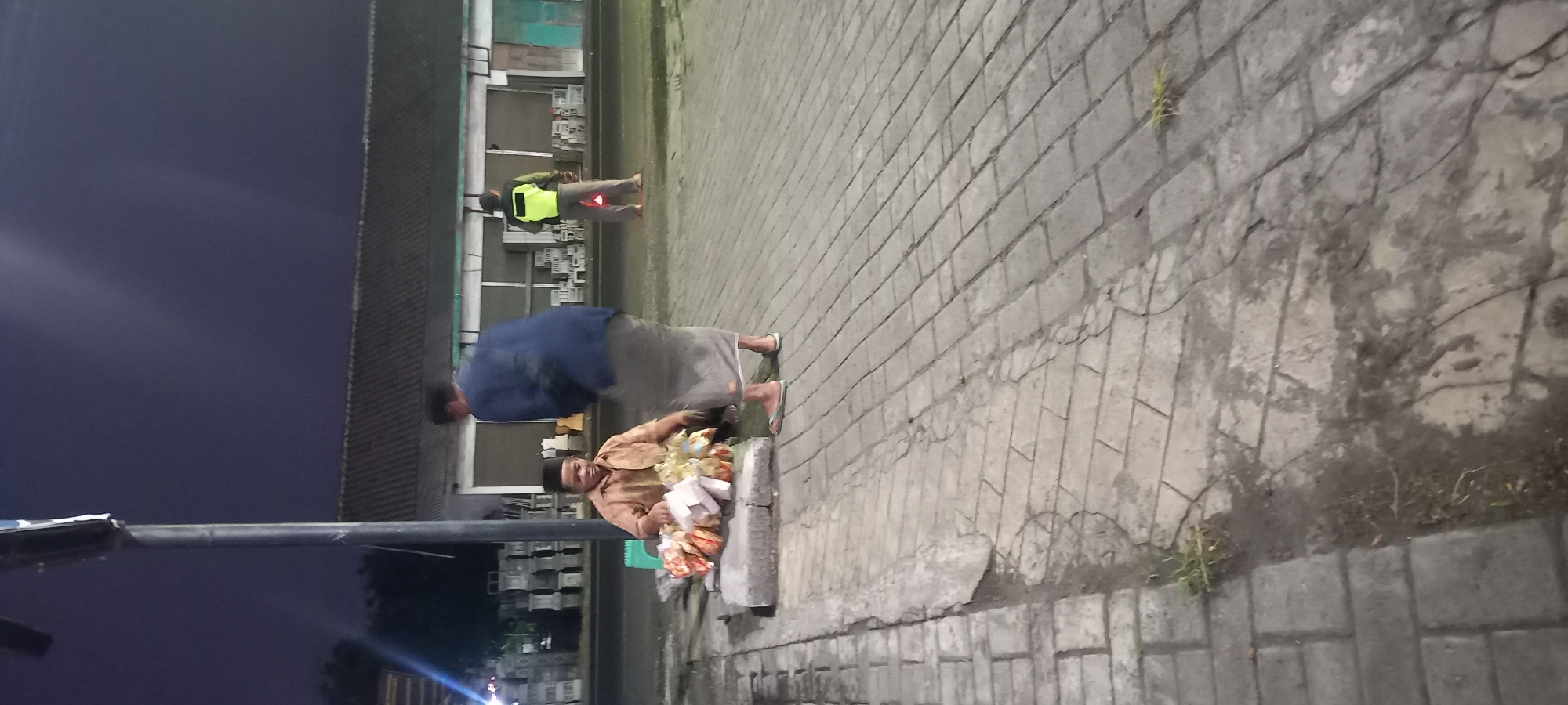 In Banjarsari, Indonesia, at night, two individuals are present on a paved sidewalk adjacent to a road. One individual, a man wearing a light brown long-sleeved shirt and a dark cap, is seated on a low concrete structure, arranging several bags of snacks. He appears to be a vendor. A second individual, a man wearing a blue long-sleeved shirt and a light-colored sarong, is standing and facing the vendor, seemingly interacting with him. In the background, there's a building with a dark, corrugated roof and what appear to be merchandise displays or construction materials outside. Further in the distance, another person is visible wearing a bright yellow vest. The sky is dark, suggesting it is nighttime. The paved area consists of rectangular interlocking stones.