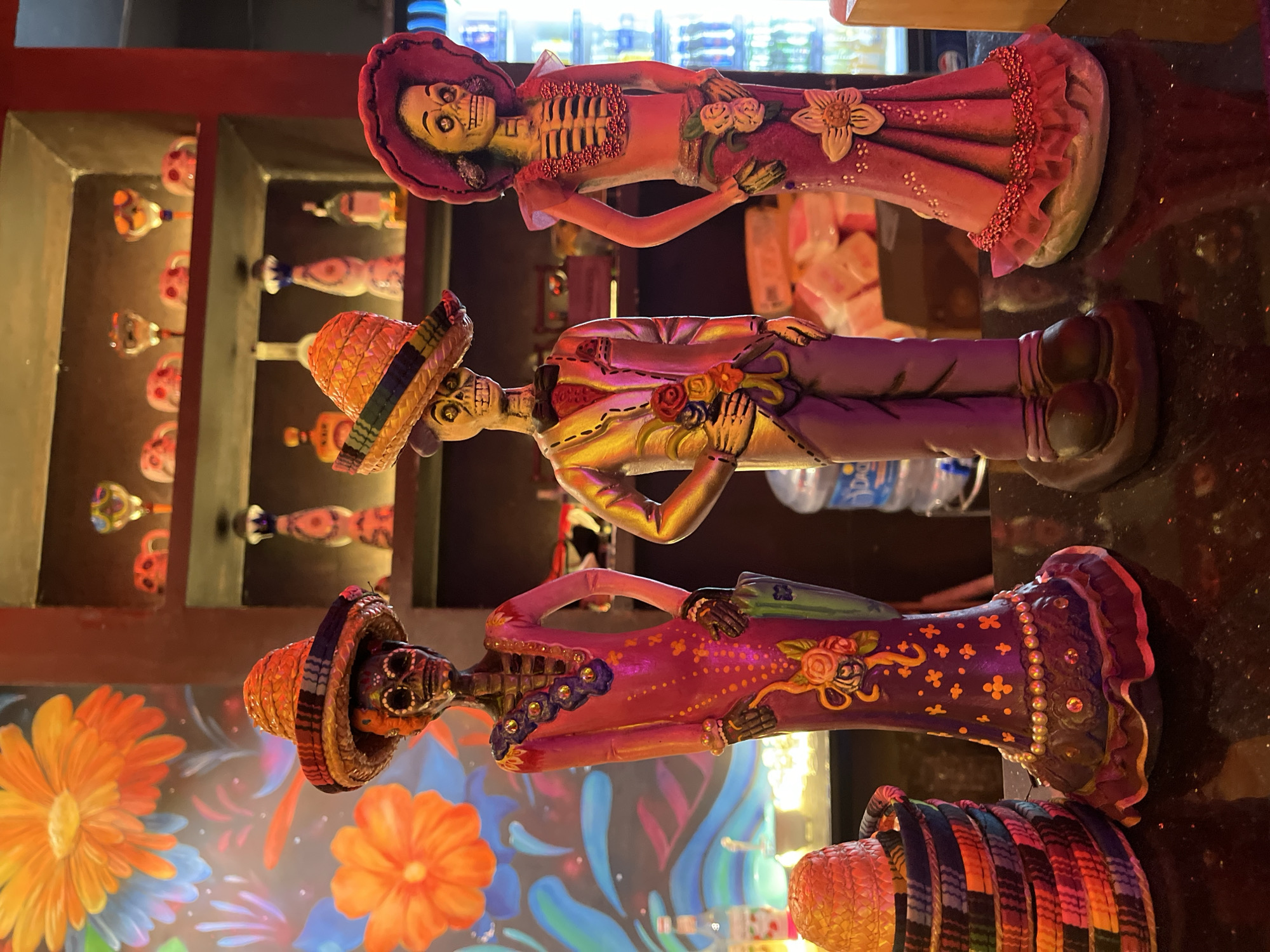 The image depicts three Day of the Dead figurines arranged vertically on a glossy, dark surface. The top figurine is a female skeleton wearing a pink dress and a wide-brimmed hat. The middle figurine is a male skeleton wearing a gold suit and a sombrero. The bottom figurine is a female skeleton wearing a purple dress and a sombrero. Behind the figurines, there is a decorated wall with floral motifs and a shelf with bottles. The location is Đà Nẵng, Vietnam, based on prior image context.
