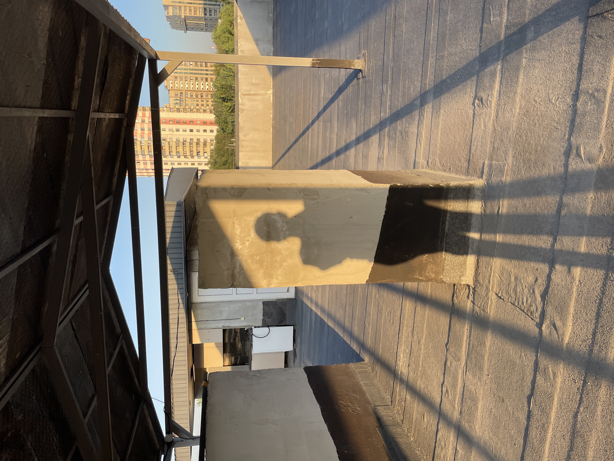The image depicts a rooftop scene in Almaty, Kazakhstan, under the warm glow of the late afternoon sun. The foreground is dominated by a flat, dark grey rooftop, marked with horizontal lines and patches of grit. Two light-colored concrete structures with dark brown bases stand prominently, casting long, sharp shadows across the rooftop. The shadow of a person, likely the photographer, is visible on the side of the central concrete pillar, indicating their presence.
In the middle ground, a light-colored metal structure with a dark grey, textured roof extends overhead, providing partial shade. Beyond this structure, more concrete walls and what appears to be an outdoor kitchen area with a white refrigerator and a black countertop are visible. A white door is set into a wall.
The background reveals a cityscape, with several multi-story residential buildings in various shades of cream, orange, and red. Lush green trees line the horizon, suggesting a moderately vegetated urban environment. The sky is a clear, pale blue, with the sun positioned to create dramatic lighting and long shadows. The overall impression is one of an everyday urban setting bathed in the golden light of sunset.