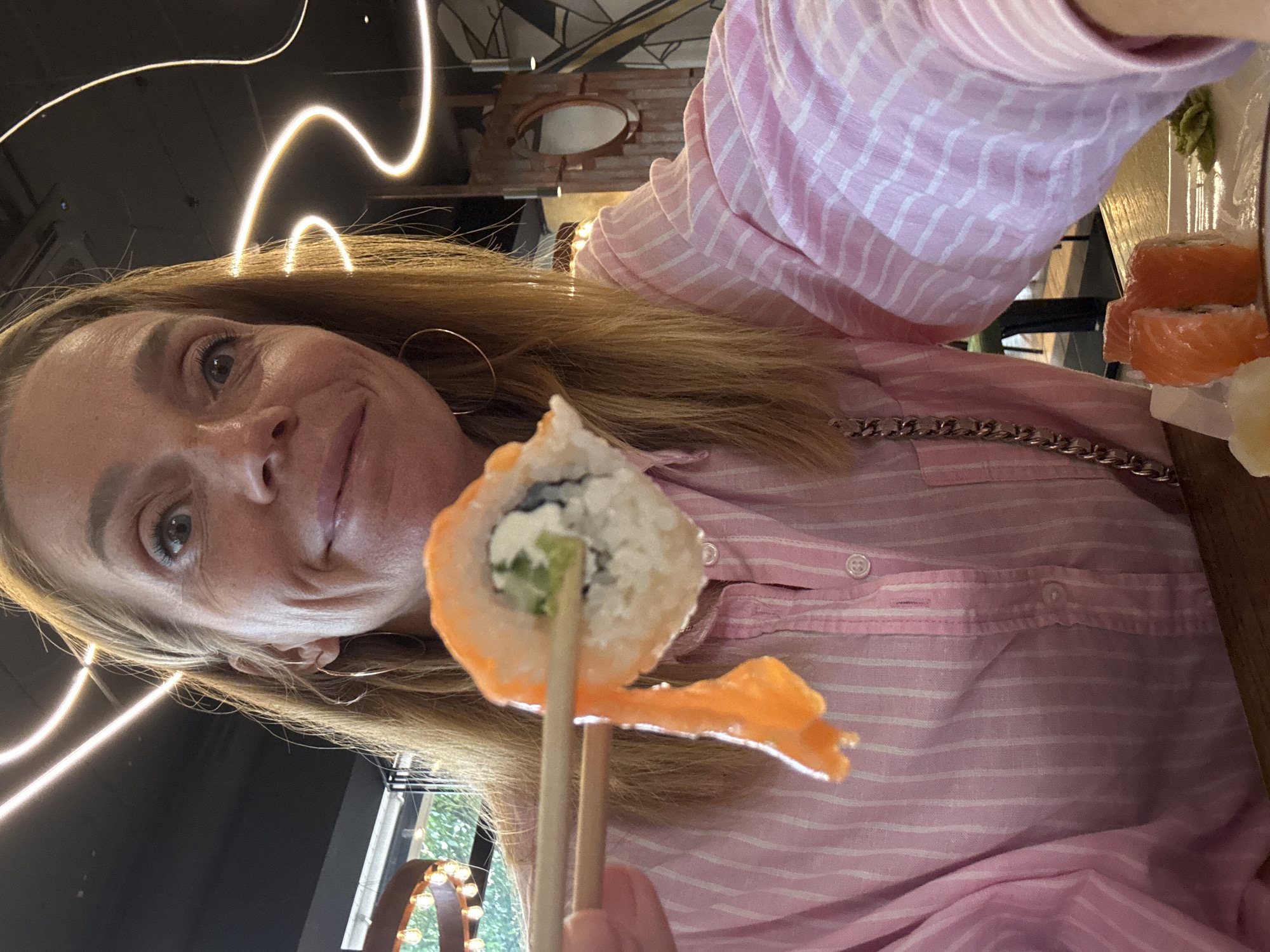 A woman with blonde hair is holding a sushi roll with chopsticks. She is wearing a pink striped shirt and hoop earrings. The sushi roll appears to have salmon, cream cheese, and cucumber. In the background, there are blurred lights and a wall with a circular mirror. The scene is likely indoors, possibly in a restaurant. The image does not contain any information about the city of Veliky Novgorod, Russia.