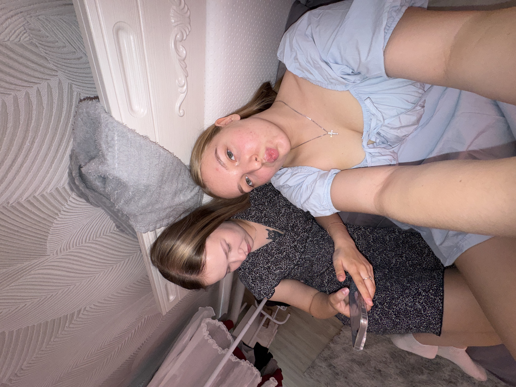 Two young women are in a bedroom. One woman is on the left, wearing a dark floral dress and holding a smartphone. She has long brown hair and a tattoo on her chest. The other woman is on the right, wearing a light blue off-the-shoulder top and a silver necklace with a cross pendant. She has blonde hair and is making a duck face. The room has textured wallpaper on the ceiling and a white headboard. A gray blanket is draped over the headboard.