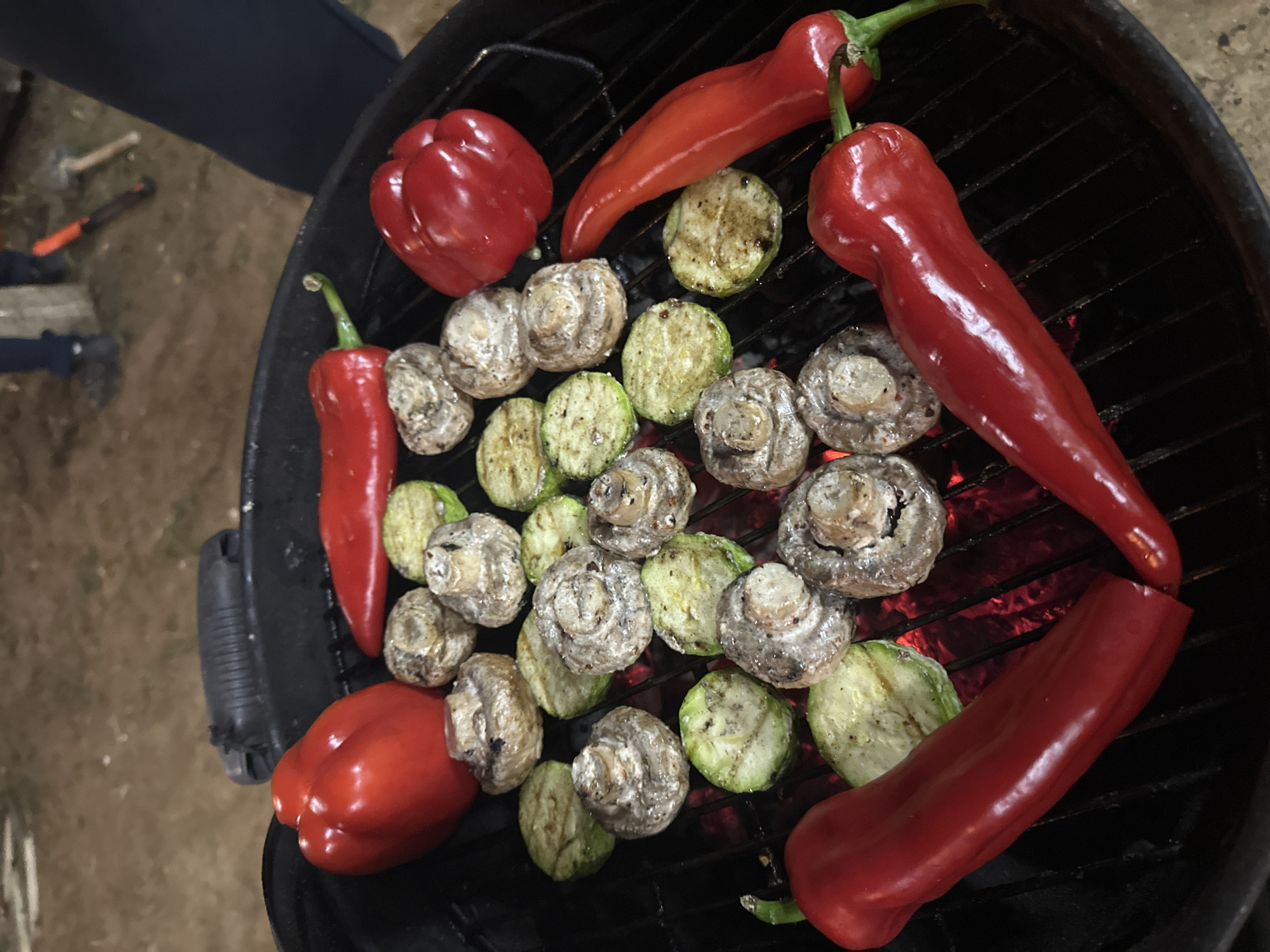 The image shows grilled vegetables on a round, black barbecue grill. The vegetables include whole red bell peppers and long, thin red chili peppers, as well as sliced zucchini and whole mushrooms. The grill is positioned outdoors, with a patch of grass or ground visible in the upper left corner. A person's leg and part of their upper body are visible in the upper left corner as well. The grilled vegetables are centered on the grill surface. The specific location is слобода Старая, Russia.