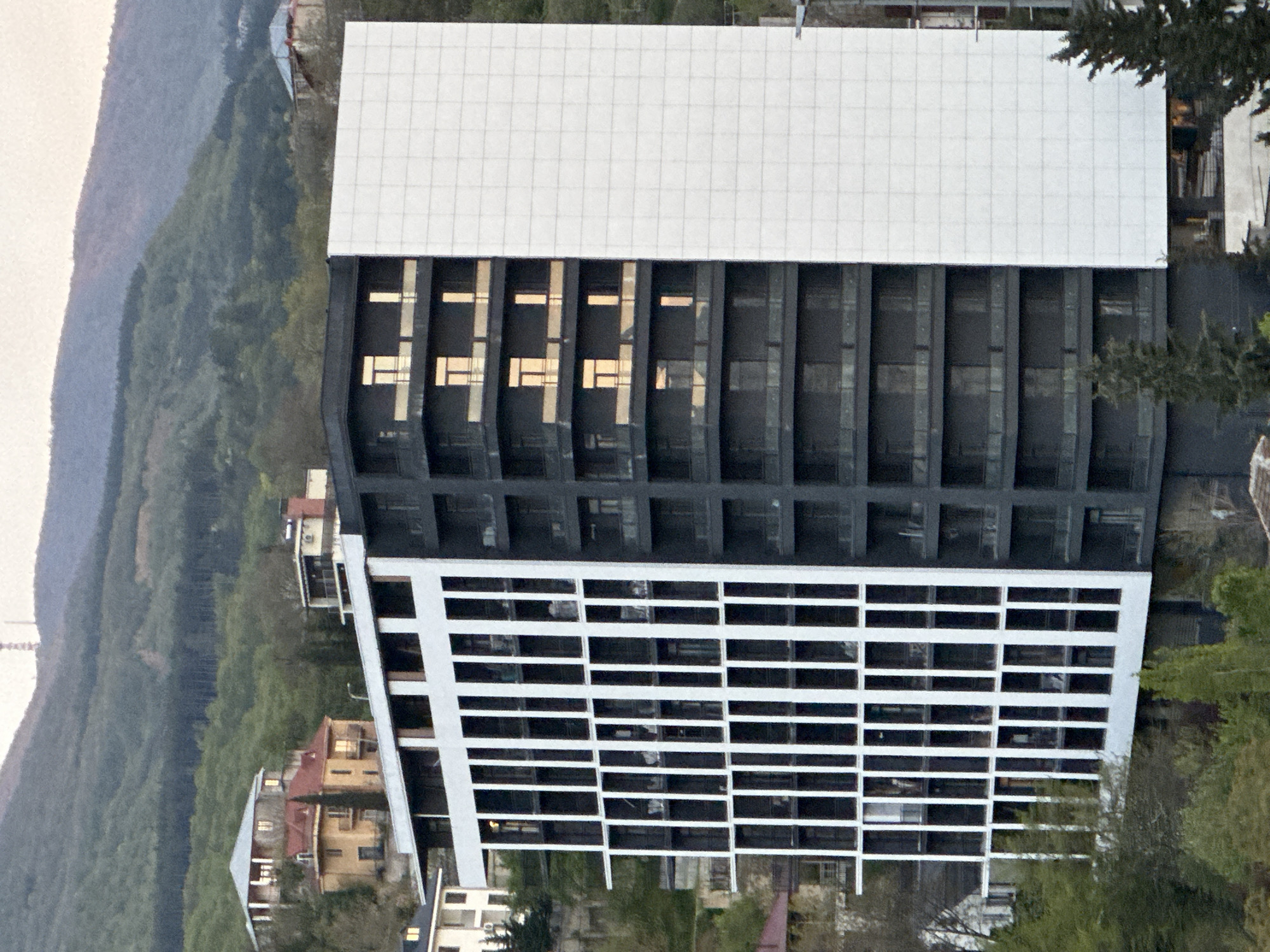 The image displays a modern, multi-story building set against a backdrop of forested hills and scattered residential houses, with the photograph itself rotated 90 degrees counter-clockwise from its natural landscape orientation.
The prominent structure is a large, contemporary building featuring distinct architectural sections. The upper portion (appearing on the right side of the rotated image) showcases a facade made of white square panels at the very top, forming a grid pattern. Below this, the building transitions to a dark, likely black or deep grey, facade characterized by deeply recessed vertical bays. Within these bays are numerous windows, many reflecting a bright, warm yellow light that forms horizontal bar-like patterns, creating a stark contrast with the dark surface.
The lower portion of the building (appearing on the left side of the rotated image) is predominantly white and features a repetitive grid of dark, rectangular window openings. This section exhibits a dynamic, angular design, appearing to subtly widen towards its base on the left side. The overall architecture is modern, with geometric lines and a strong contrast between white and dark tones.
In the background, behind the large building, are lush green, forested hills or mountains, indicative of a natural, elevated landscape. Scattered among the trees and below the main building are glimpses of smaller, more traditional residential houses with pitched roofs, some featuring earthy colors like yellow and red. The lighting suggests a soft, diffused light, possibly from an overcast sky or during the early morning/late afternoon, as indicated by the warm reflections in the windows. No text is visible in the image. The setting suggests an urban environment nestled within a natural, hilly terrain, consistent with Tbilisi, Georgia.