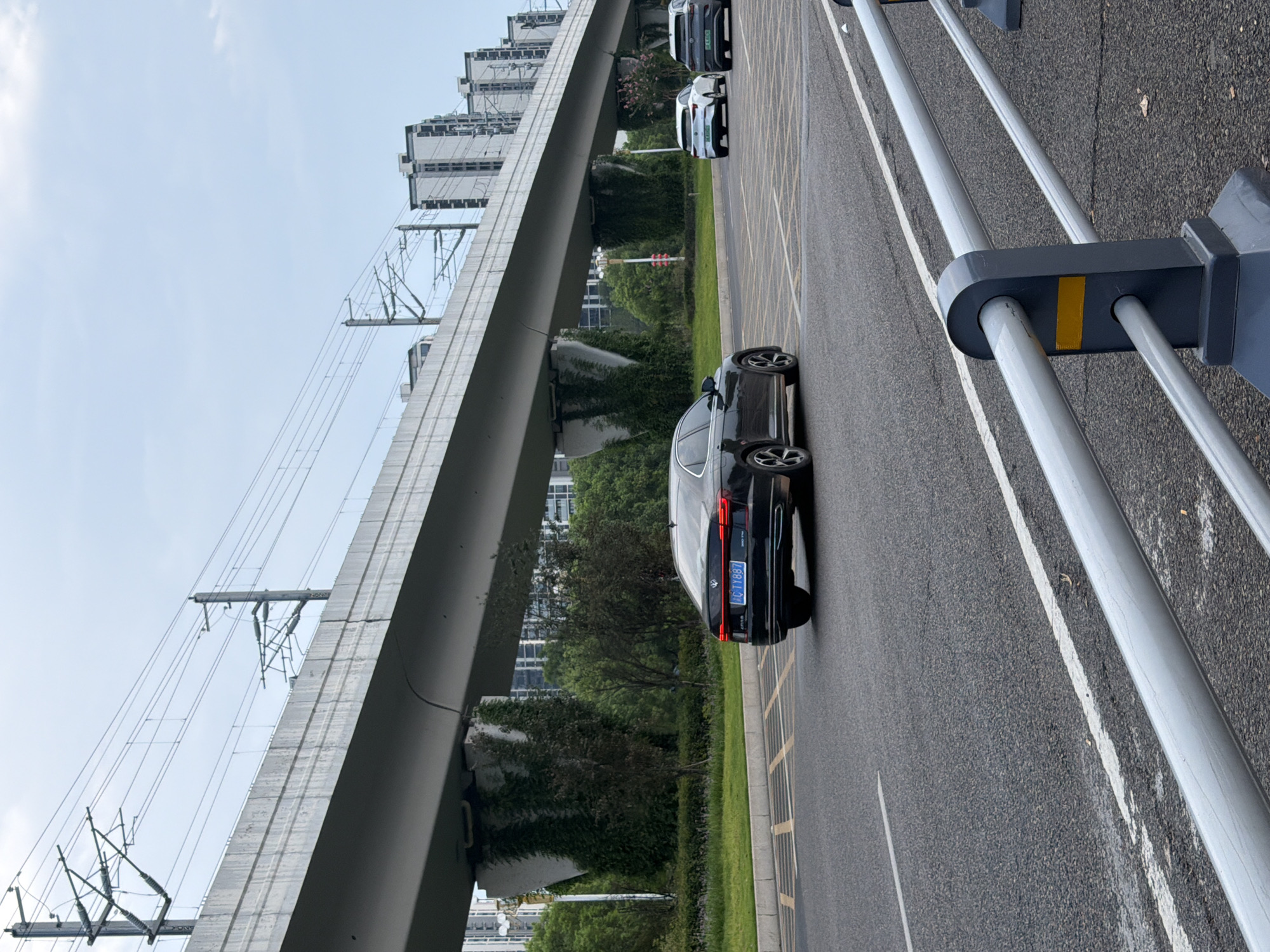 This image captures a view of an elevated transportation infrastructure, likely a highway or railway overpass, in the city of Jiangjin, China. The overpass is supported by concrete pillars that are partially covered in green ivy. In the foreground, a black Volkswagen sedan is driving on a multi-lane asphalt road. The car's license plate is visible and reads "C·TY887". Further in the distance, two other vehicles are visible on the road: a white car and a dark gray SUV. The background shows a cluster of modern, high-rise residential buildings. The sky is overcast with some clouds. The scene appears to be an urban environment with ongoing traffic and infrastructure development.