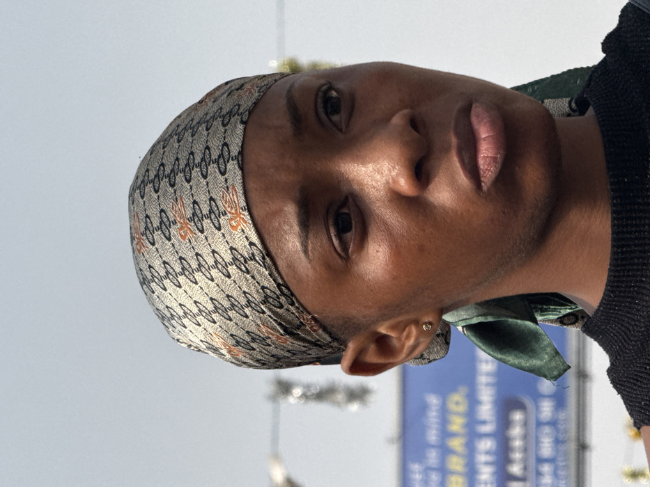 A close-up, chest-up outdoor portrait depicts a young adult Black female in Lekki, Nigeria, positioned slightly right of center. She has a dark complexion, dark eyes, and a neutral facial expression with slightly parted lips. Her head is covered by a patterned headscarf featuring a light tan background with repeating black chain-link patterns incorporating 'G' shapes and intermittent orange bird-like motifs. A dark green fabric tie from the scarf is visible behind her left earlobe, which contains a small, circular, silver-colored stud earring. She wears a dark, possibly black, textured top. The background consists of a light, hazy sky. Behind her left shoulder, a blurred blue billboard or sign is partially visible, displaying white text including "in mind," "BRAND," "MENTS LIMITED," and "Lekki Akoka," confirming the urban location within Lekki, Nigeria. Additional indistinct, reflective elements are present as bokeh in the far background.