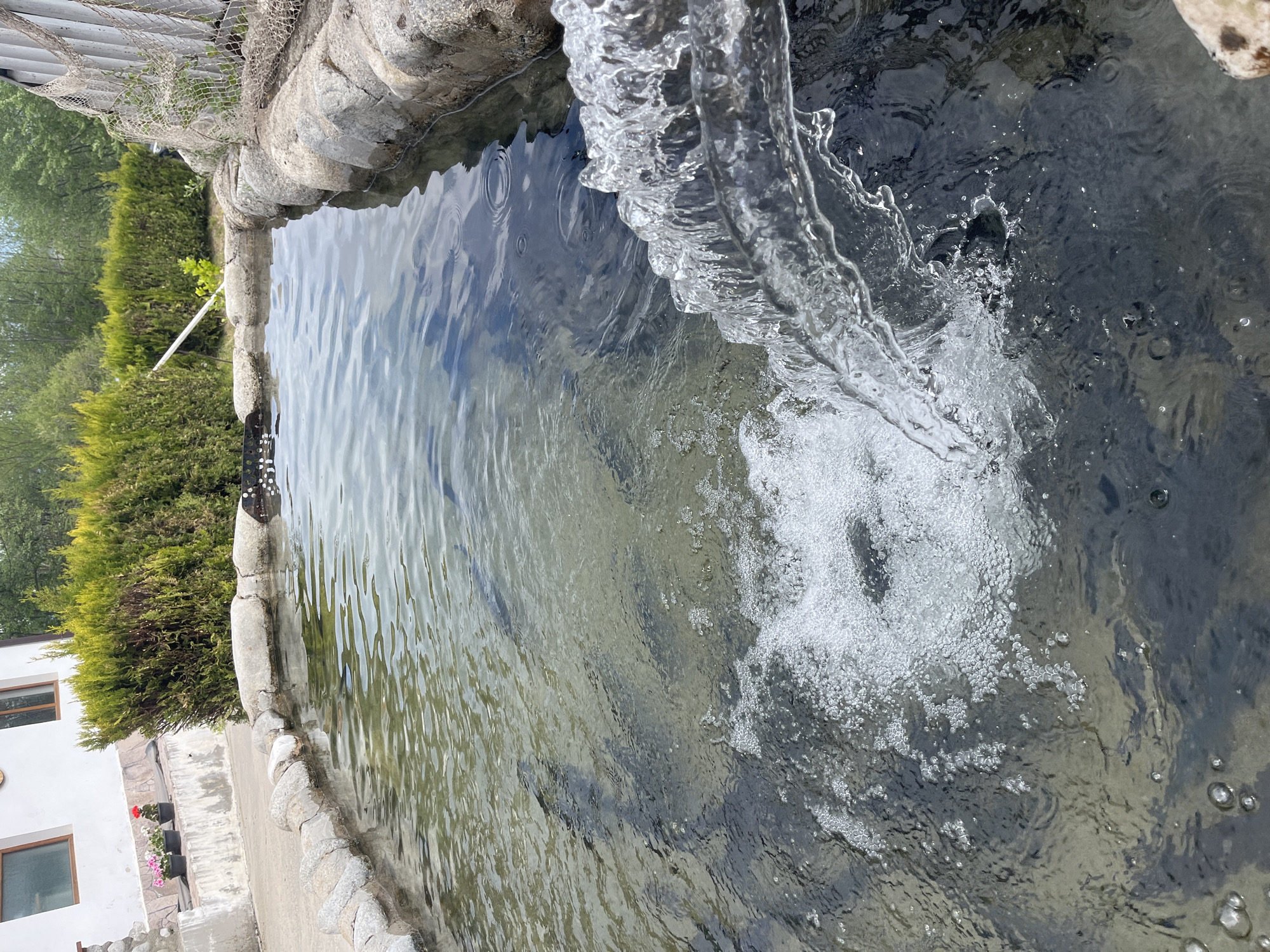 The image shows a water feature, possibly a fountain or a small pond. Water is actively pouring into the pool, creating splashes and bubbles. The pool is surrounded by rocks and concrete forming a border. In the background, there is greenery and part of a building with windows. The location is Unknown, Unknown.