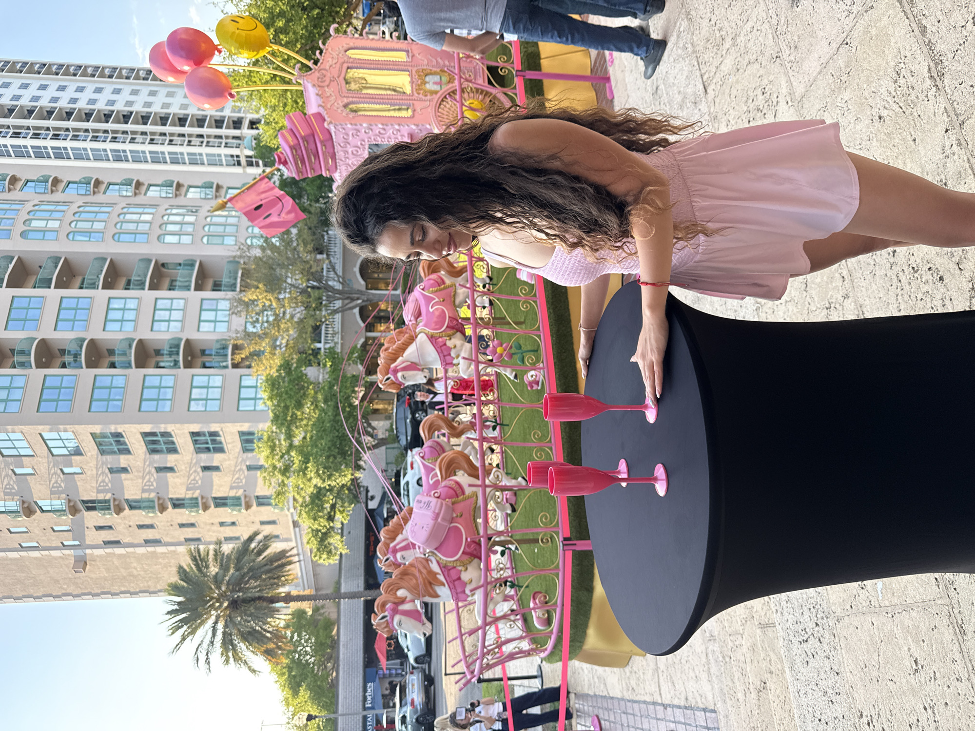 An outdoor, eye-level, wide-angle shot depicts an urban scene focused on a young adult female standing beside a tall, black, cylindrical cocktail table. The female, positioned right of center, has long, wavy, dark brown hair, light skin, and wears a light pink strapless top and matching skirt. She rests her hands on the table, looking down and to her left with a subtle smile. A red beaded bracelet adorns her left wrist. On the black table are three pink champagne flutes, two upright and one slightly toppled.
Behind the female and the table is a large, ornate, pink and gold display featuring multiple carousel-style horses with orange manes and tails, arranged on a raised platform with artificial green turf and encircled by a pink railing. A pink carriage-like structure, adorned with stacked pink boxes and a pink flag bearing a smiley face, stands on the right side of the display, along with three balloons (two pink, one yellow smiley face). Pink ribbon barriers delineate the display area.
In the mid-ground, other individuals are visible: a person in a gray t-shirt and blue jeans partially visible on the far right, and another person holding a camera near the left pink barrier. Further back, vehicles are parked on a street.
The background features multiple modern, light-colored high-rise buildings with numerous balconies and teal-tinted windows. A prominent palm tree is visible on the left. The distinct "COASTAL" and "Forbes" signage is visible on a building in the background. This urban environment, characterized by modern architecture, palm trees, and event signage, specifically identifies the location as downtown Saint Petersburg, Florida, United States of America, likely near Central Avenue. The scene is illuminated by bright daylight, suggesting late morning or afternoon.