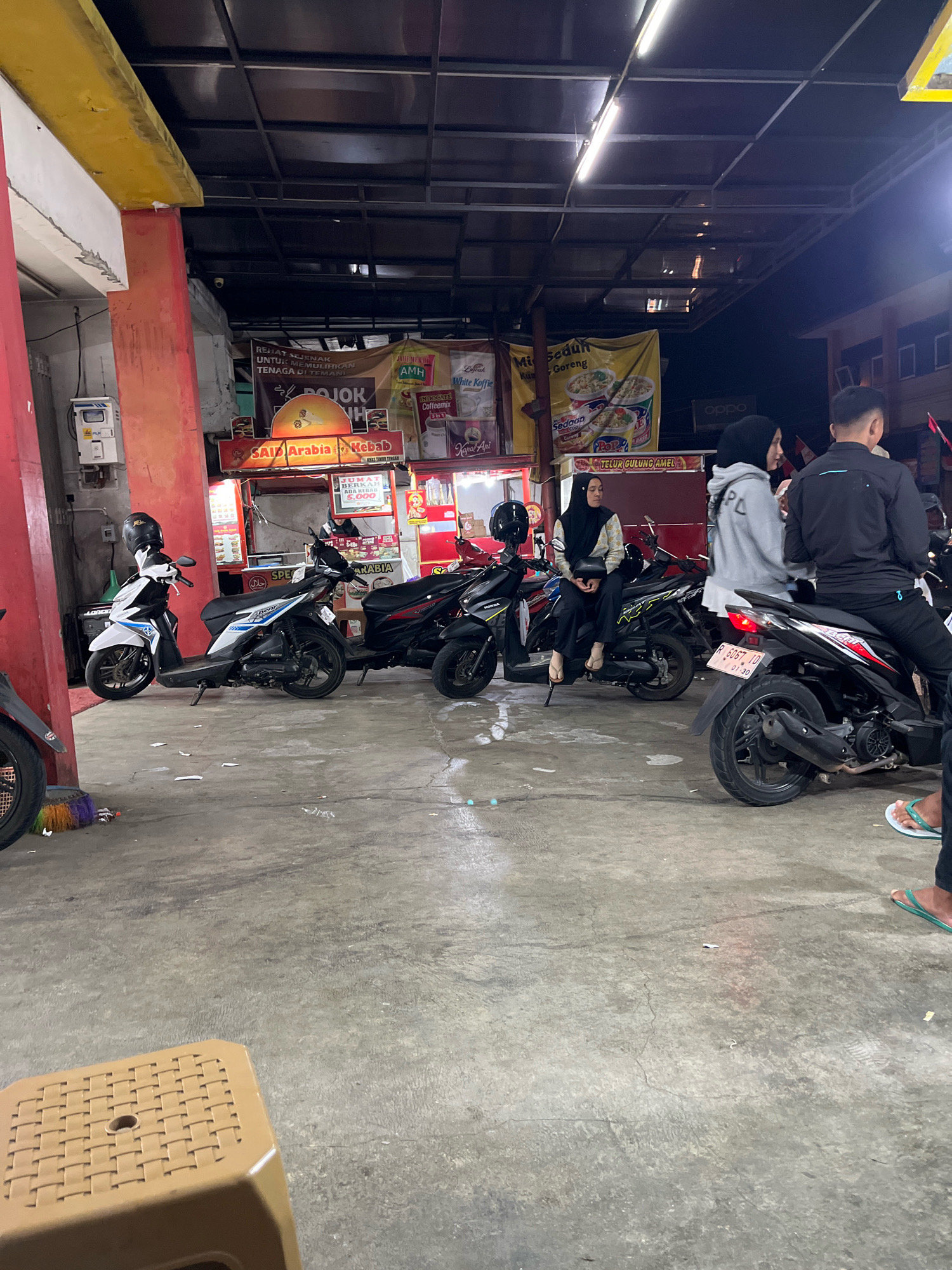 The image depicts an outdoor scene at night, likely in Karangkobar, Indonesia, where several motorcycles are parked. In the foreground, a light brown woven plastic stool is positioned on a concrete floor. The middle ground features a row of parked motorcycles, some with riders. A woman in a black hijab and patterned top is seated on one motorcycle, while another woman in a grey hoodie and black hijab stands next to a motorcycle. A man in a black jacket is also visible standing near a motorcycle. Behind the motorcycles, there are food stalls or small eateries with banners and signs, suggesting a bustling night market or street food area. The stalls display various products and advertisements, including "SAID ARABIA KEBAB" and banners with text in Indonesian. The environment is dimly lit, with overhead lighting from the stalls and possibly streetlights. The concrete floor shows signs of wear and some debris.