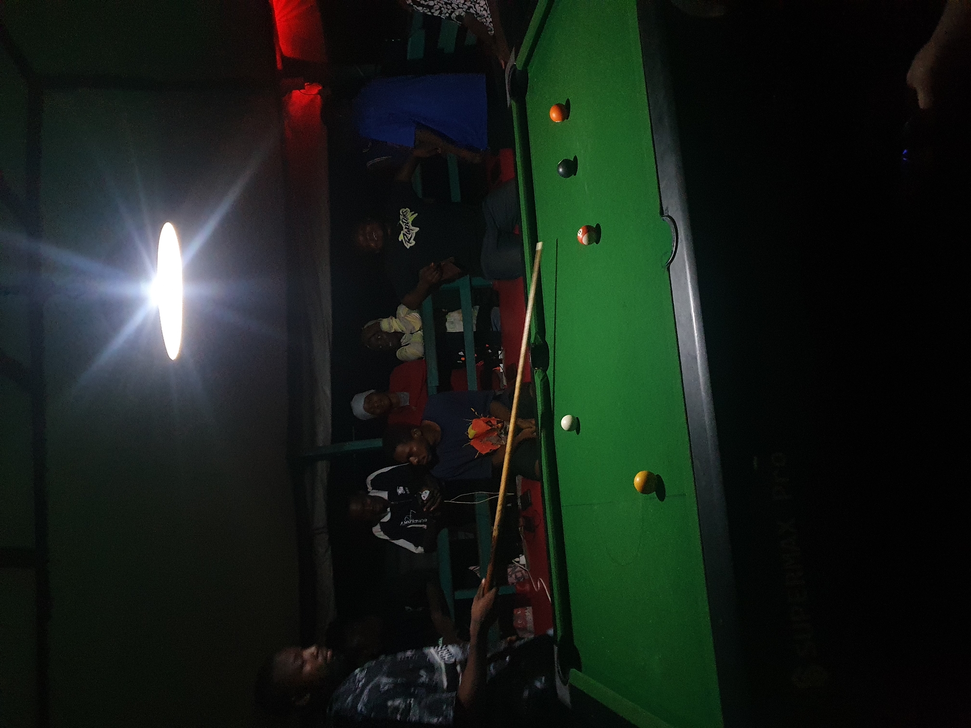 A group of people are gathered around a pool table in an indoor setting, possibly a bar or recreational area, in Lokoja, Nigeria. The lighting is dim, with a bright overhead lamp illuminating the pool table and casting shadows. The pool table is green, with several balls on it: a white cue ball, a yellow ball, a black ball, and an orange ball. A man is holding a pool cue, appearing to be in the process of playing. Other individuals are standing or sitting in the background, some watching the game. The overall atmosphere seems relaxed and social. The environment appears to be a covered outdoor space or a room with a low ceiling, with some red lighting visible in the background. The pool table has "SUPERMAX Pro" branding on its side.
