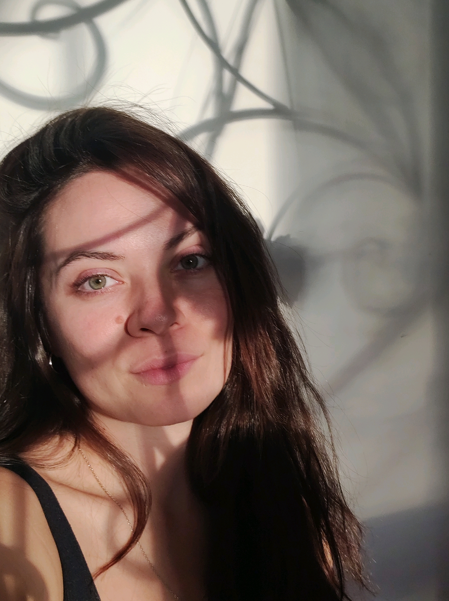 In a dimly lit indoor environment, a young Caucasian woman is captured in a close-up, head-and-shoulders portrait. Her long, dark brown hair is styled loosely around her face. She is wearing a black tank top, and a delicate gold chain necklace is visible around her neck. Her face is turned slightly to her left, and her gaze is directed forward. She has light-colored eyes and a light complexion, with a subtle mole visible on her right cheek. A shadow, cast by an unseen object, falls diagonally across her face, obscuring part of her forehead and cheek. The background appears to be a wall with abstract shadow patterns, possibly from a nearby window or light source. The image does not contain any specific landmarks or contextual clues that would allow for the determination of its location within the city of Фоки, Russia.
