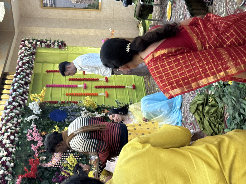 The image captures an indoor ceremonial gathering in Malasar, India. The primary background consists of a lime green fabric wall extensively decorated with artificial green foliage. An archway of white and maroon artificial flower garlands spans the top, with vertical garlands of white, orange, pink, and red flowers suspended below. A decorative crescent moon motif with dangling elements and a blue circular woven ornament are affixed to the green backdrop. The floor is covered with a patterned carpet, overlaid with scattered green artificial leaves and a crumpled green cloth in the foreground.
Five individuals are clearly visible, with partial views of others. In the mid-ground, a young woman is seated on the floor, facing slightly left. She wears a yellow and light blue ombre saree embellished with silver sequins, a necklace, earrings, and a maang tikka. Her dark hair is styled with a center part. Standing directly behind her, with his back mostly to the camera, is a man in a horizontal striped sweater (maroon, black, white) and a brown scarf, holding a round silver tray with multiple small metallic bowls containing ceremonial items. His lower body is covered by a patterned yellow garment. To the right of the seated woman, a man stands facing left, wearing a light-colored patterned shirt and dark trousers.
In the immediate foreground, with their backs to the camera, are two individuals: one on the left in a solid yellow kurta, and one on the right, a woman in a red saree with a gold checkered pattern and a red blouse. Her dark hair is pulled into a ponytail secured with a silver decorative clip. On the far right, a black video camera on a light-colored tripod is positioned, indicating recording of the event. A textured light brown wall with a framed landscape picture is visible to the right of the main green backdrop.