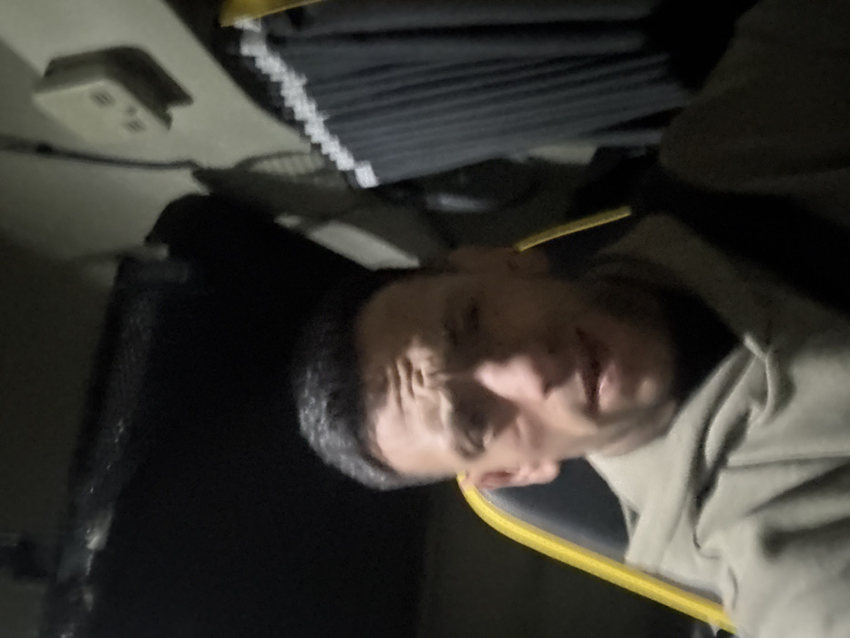 This image depicts a single male subject positioned centrally within what appears to be the dimly lit cabin of a commercial truck, located in Foggia, Italy. The man, with short, dark hair and a visibly furrowed brow, looks directly towards the viewer. His face exhibits a slight stubble and a subtle reddish mark or scar on his left cheekbone. He is dressed in a light olive green hooded sweatshirt, with a dark seatbelt strap extending diagonally across his chest and right shoulder. He is seated in a dark vehicle seat featuring a prominent yellow or bright green trim along its left side. The interior environment behind him includes a dark overhead storage unit or bunk. To the right, a light-colored wall panel hosts a small, off-white electrical box with two circular inputs. Below this, dark, pleated privacy curtains with a light-colored upper trim are drawn. A dark, circular ventilation fan or vent is partially visible in the upper right quadrant. The scene is illuminated by low-level ambient light, with the subject's face appearing more brightly lit than the background.