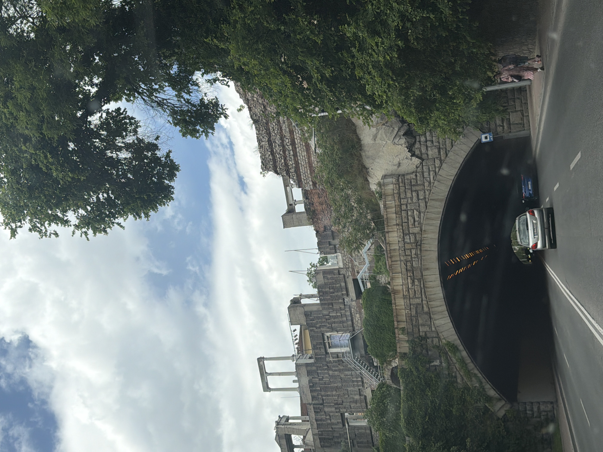 The image depicts a tunnel entrance in Plovdiv, Bulgaria. A car is partially inside the tunnel. The architecture above and next to the tunnel includes stone and brickwork, along with vegetation. Trees are visible in the upper portion of the image, obscuring the sky. The day appears to be overcast, with cloudy skies. A road runs alongside the tunnel.