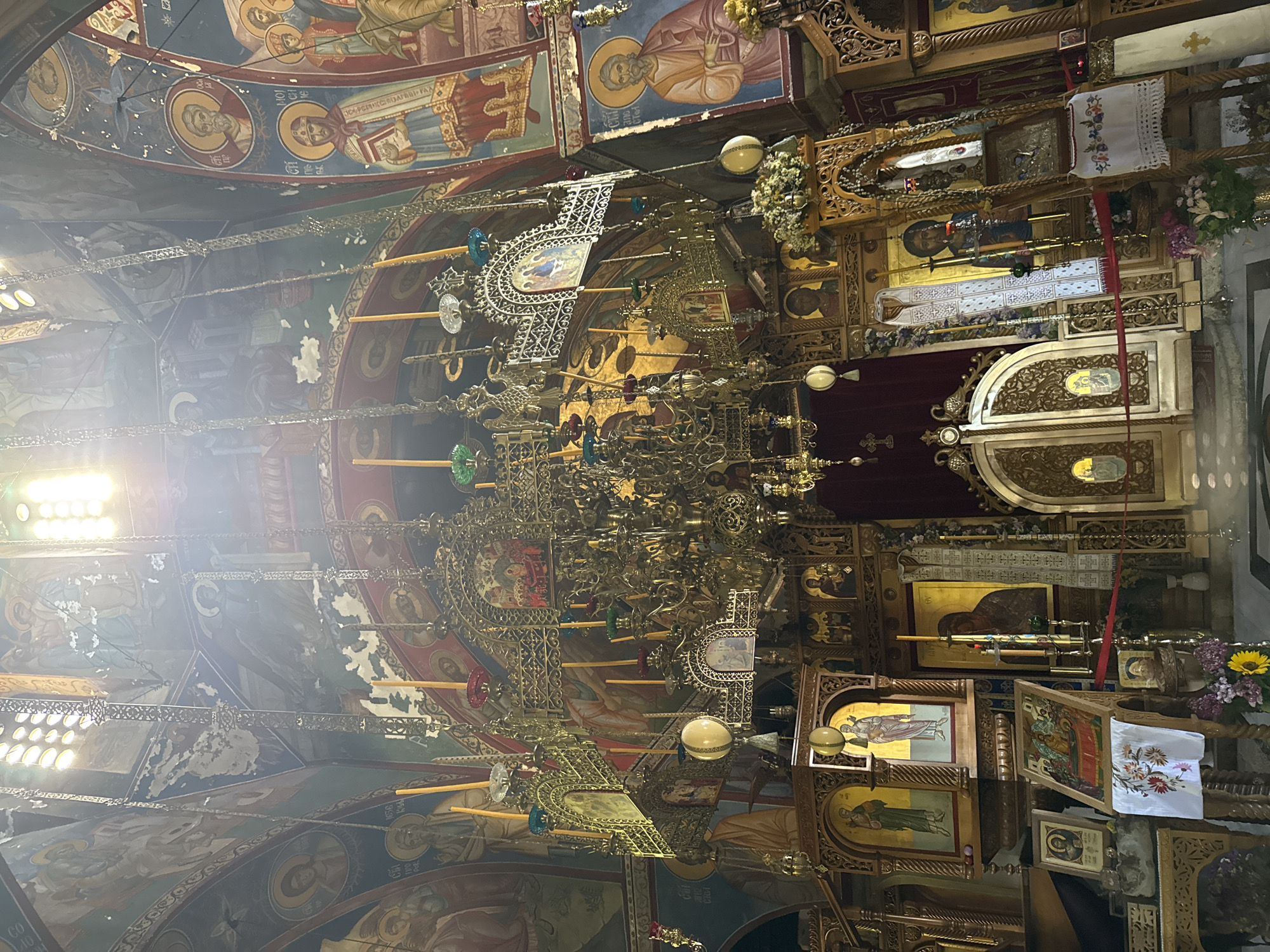 The image captures the interior of a church in Trebinje, Bosnia and Herzegovina, likely the Church of the Transfiguration of the Lord, given the ornate Byzantine style and iconostasis visible. The perspective is from a low angle, looking upwards towards the elaborately decorated ceiling and a large, golden chandelier adorned with numerous candles. The walls are covered with religious frescoes depicting saints and biblical scenes. The lower portion of the image shows a richly carved wooden iconostasis, a screen separating the nave from the sanctuary, featuring icons of religious figures. Incense burners, candles, and embroidered cloths are also visible, contributing to the sacred atmosphere. The lighting appears to be a mix of natural light filtering in from above and the ambient light within the church. There are no people prominently featured in the scene.