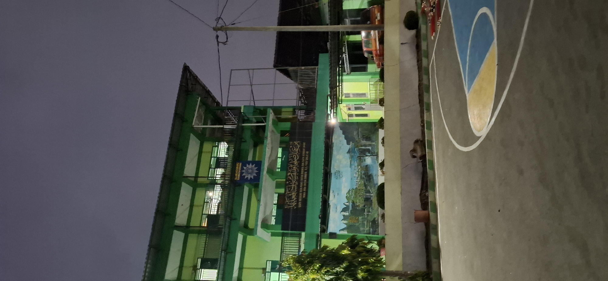 The image depicts a multi-story building with a predominantly green facade, situated in what appears to be an outdoor court area. The building has multiple levels with balconies and windows, some of which are illuminated from within. A large mural or banner featuring Arabic calligraphy and a symbol, possibly a logo or insignia, is prominently displayed on the facade of the building. Below this, a colorful mural depicting a landscape with water, mountains, and trees is visible. A small orange bucket is placed near the base of the building. In the foreground, a section of a basketball court with blue, yellow, and white markings is visible. To the right of the building, a portion of a red vehicle is parked. The sky above is dark, suggesting it is either dusk or nighttime. The overall scene appears to be in a public or institutional setting, possibly a school or religious institution, given the calligraphy and the mural's themes. The location is identified as Wonorejo, Indonesia.