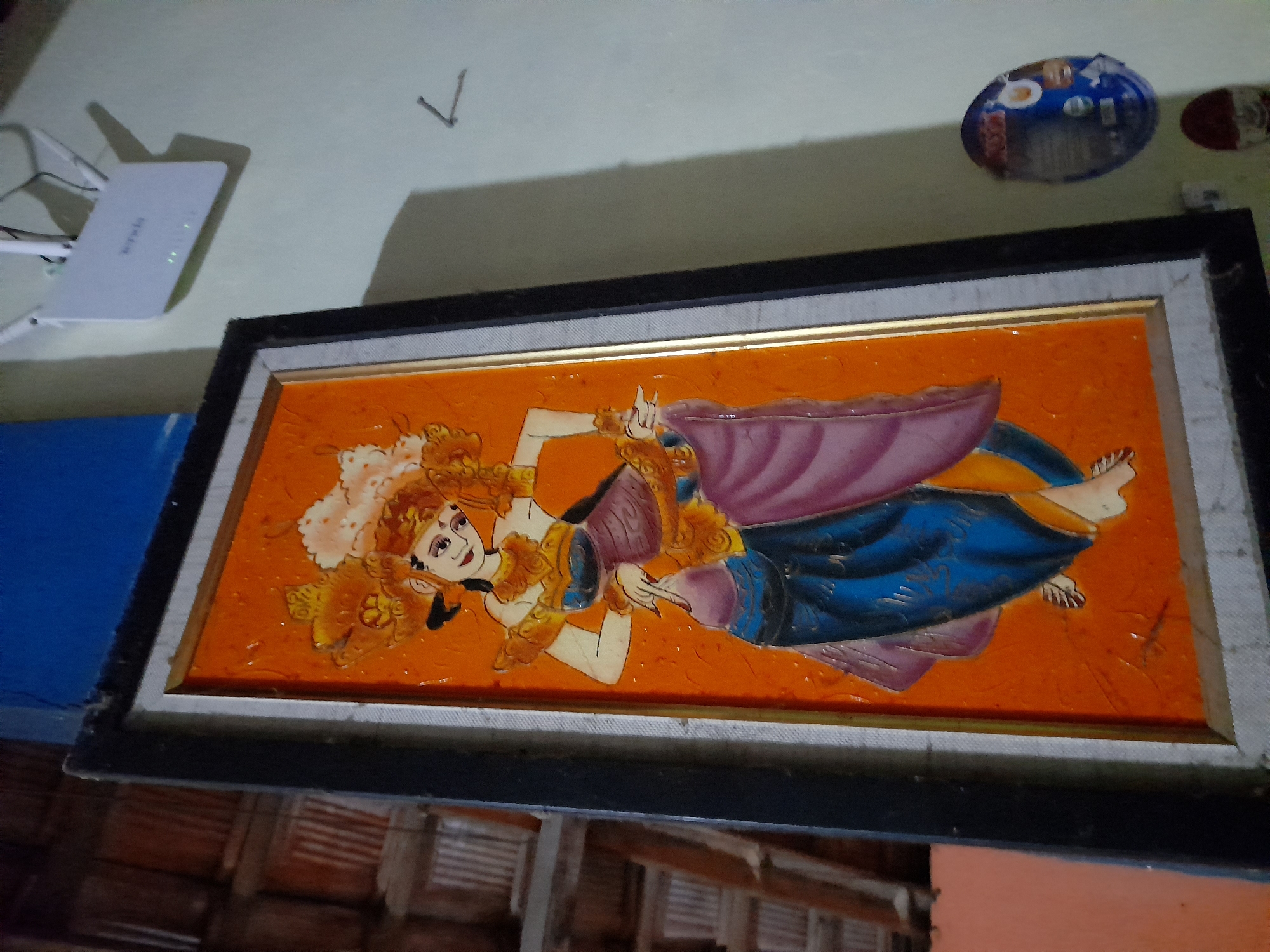 The image depicts a framed painting of a woman in traditional attire. The painting is horizontally oriented with an orange background and features a woman with elaborate, reddish-orange hair. She is depicted in a reclining or dancing pose, wearing a blue and purple garment adorned with gold accents. Her skin is fair, and she has dark eyes and lips. The painting appears to be made of some textured material, possibly with a glossy finish. The frame is dark and has a lighter inner border.
Below the painting, on the wall, is a small, circular object that appears to be a lid or a plate with various items on it, possibly food or spices. To the right of the painting, a white Wi-Fi extender is mounted on the wall, indicated by its shape and the visible indicator lights. The wall itself is painted a light, neutral color. The overall impression is of an indoor setting, likely a domestic or display area, in a residential context. The reference to Pulokulon, Indonesia suggests a potential location for this scene, though the image itself does not contain explicit geographical markers. The background above the painting shows wooden ceiling beams, typical of traditional Indonesian architecture.