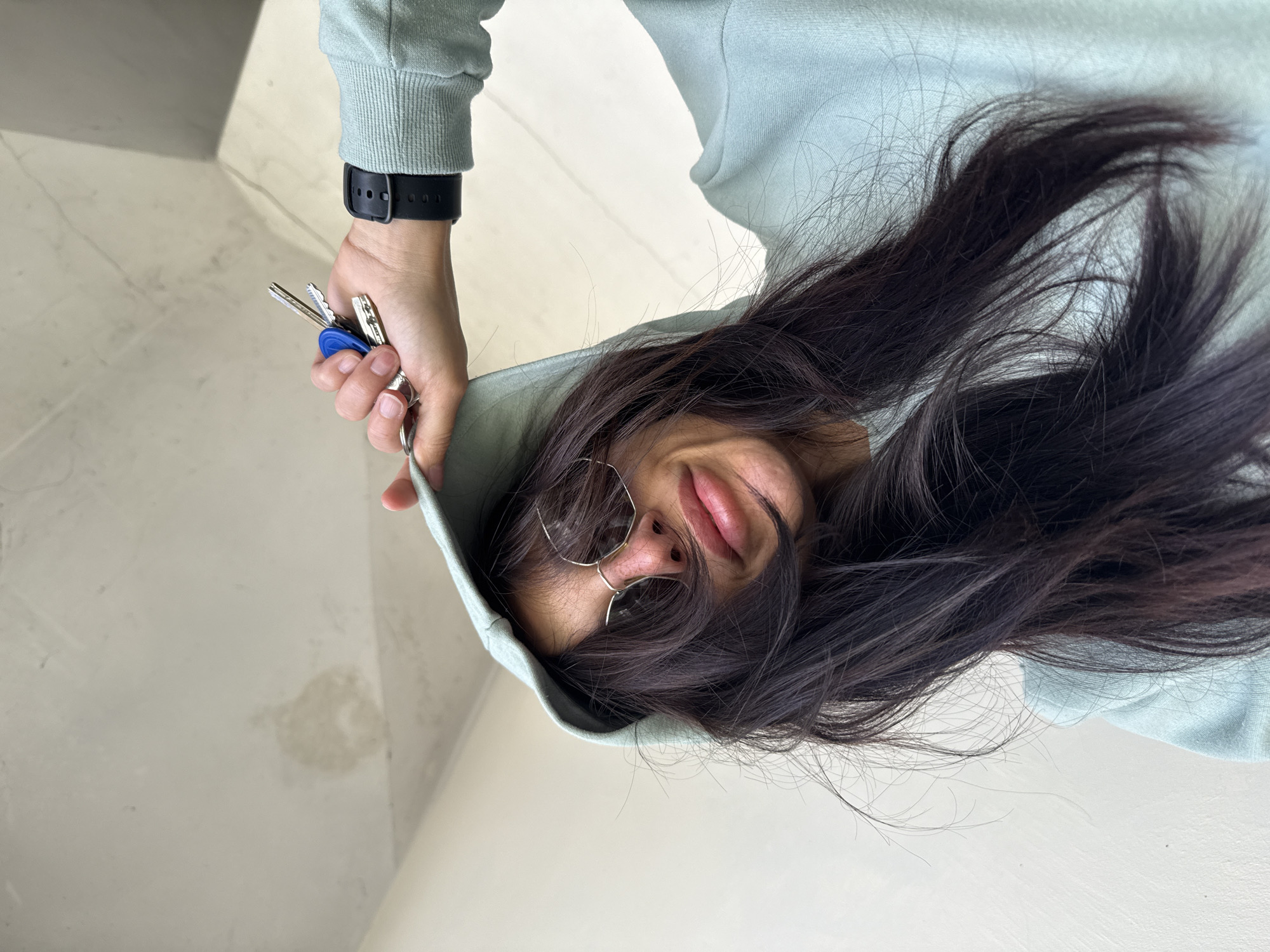 A young woman with dark hair and glasses, wearing a light blue hooded sweatshirt, is holding a set of keys. She is looking downward. Her wrist shows a black smartwatch. The background consists of a white wall and ceiling. The location in Pleven, Bulgaria, cannot be determined from the image.