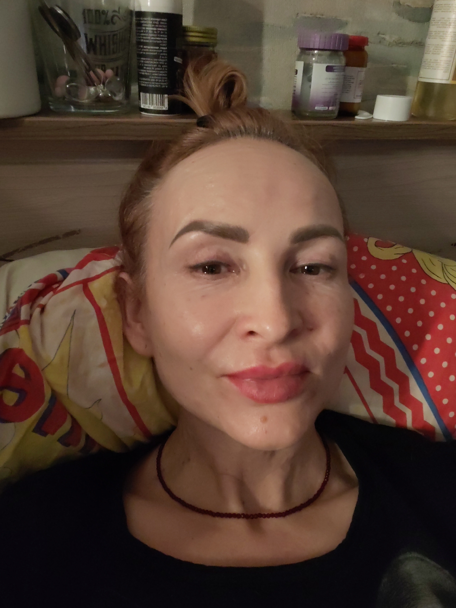 A woman is lying down, possibly on a bed, with a colorful pillow behind her head. She has her hair in a high bun. She appears to have fair skin and is wearing a dark top and a beaded necklace. The background includes a shelf with various bottles, jars, and a glass containing utensils. The location is Chaikovsky, Russia.