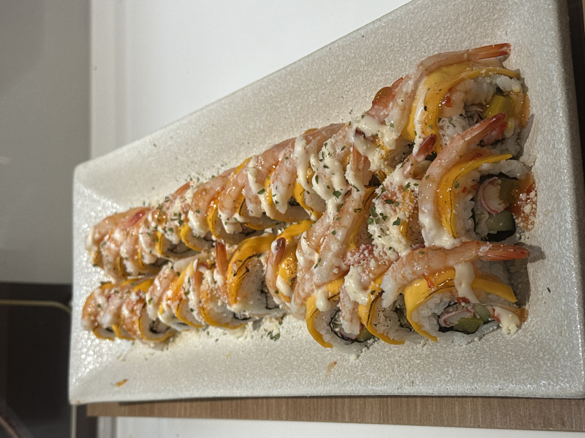 A platter of shrimp sushi rolls is displayed on a white, textured ceramic plate. The sushi rolls are arranged in a diagonal line, from the top left to the bottom right of the image. Each roll appears to be wrapped in rice and nori, with a bright orange filling that resembles mango. Cooked shrimp are draped over the top of each roll, and a creamy white sauce is drizzled over the shrimp and mango. Finely chopped green herbs are sprinkled over the top of the sushi. The platter is placed on a white countertop, with a hint of a wooden surface on the left side of the frame. The background is out of focus, suggesting an indoor setting. This image was taken in Nha Trang Ward, Vietnam.