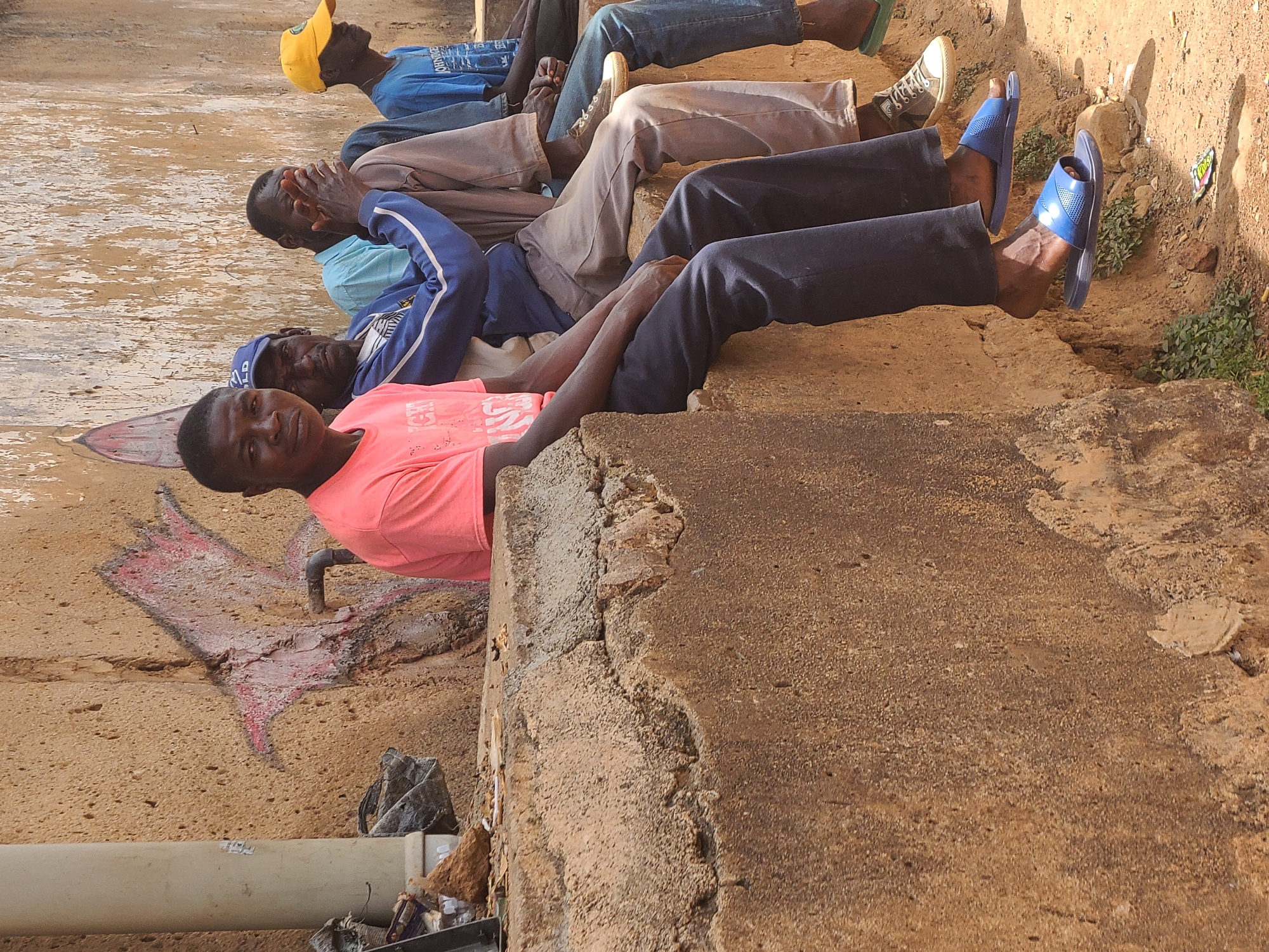In an outdoor setting in Jos, Nigeria, five men are lying down, apparently resting. The man closest to the camera wears a coral-colored shirt and dark pants, and is positioned on a concrete ledge near a wall with faded paint. Behind him are men in a blue jacket, a tan shirt, and blue shirts, one with a yellow hat. The men are wearing casual shoes. The ground around them is sandy and dotted with vegetation. The ledge where the closest man is resting is a large, roughly textured concrete structure. Visible pipes and miscellaneous debris are near the wall.
