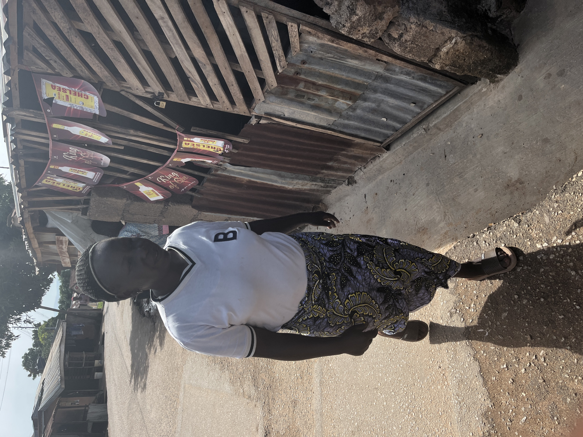 A medium shot captures a woman walking on a dirt road in a dense urban settlement in Jos, Nigeria. The woman is wearing a white v-neck t-shirt with a black letter "B" logo on the left side of her chest and a long, patterned skirt. She has dark skin, is wearing a black headband, and is wearing brown sandals. Behind her, a roadside structure is visible, constructed from corrugated metal and wooden planks. A string of red banners, featuring images of bottles and text, is draped across the front of the structure. The environment appears dusty and underdeveloped, with other structures and trees visible in the background. The lighting suggests it is daytime, with strong shadows cast by the woman and the structures.