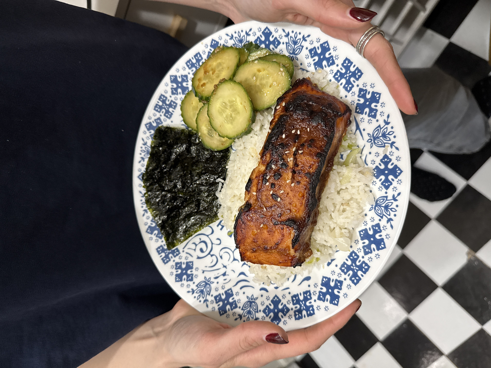 The image shows a person holding a white plate with blue decorations, containing a meal consisting of cooked salmon, rice, seaweed, and sliced cucumbers. The person is wearing a dark blue dress or shirt and has a ring on their finger with maroon nail polish. The background is a black and white checkered floor. This image likely does not contain specific information that is specific to New York, United States of America.