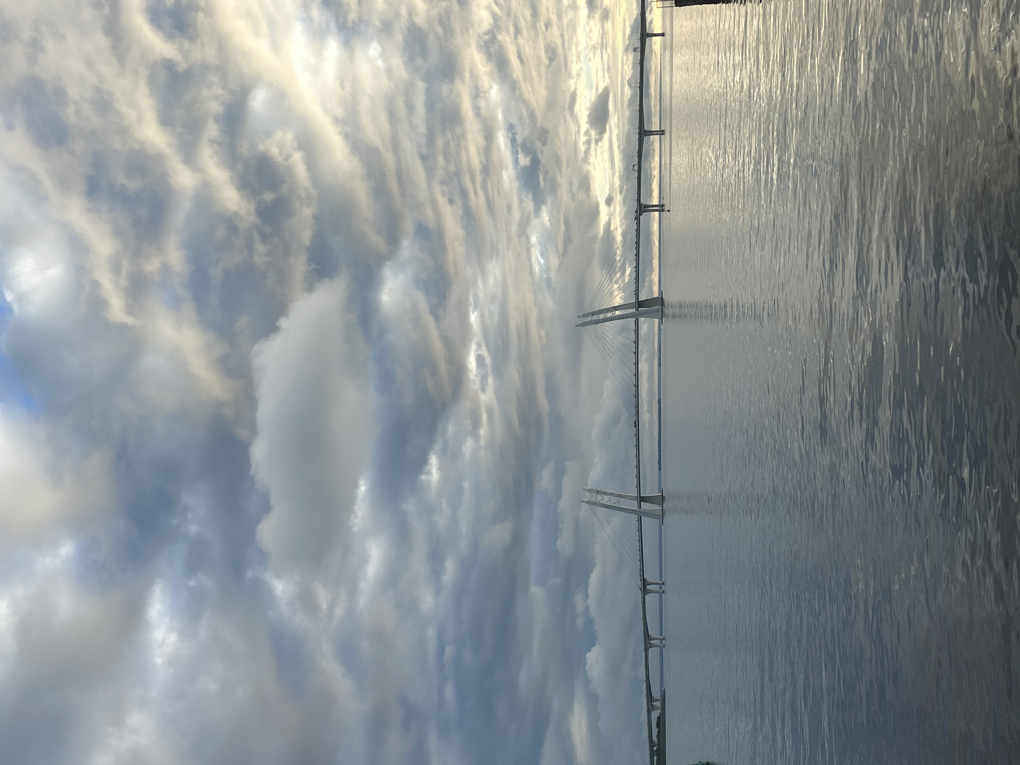 A wide-angle, eye-level, outdoor shot captures a cable-stayed bridge spanning across a body of water. The bridge, with its distinct white pylons and network of cables, stretches horizontally across the frame. Vehicles are visible on the bridge, indicating it is in use. The water's surface is calm with gentle ripples, reflecting the overcast sky. The sky itself is filled with a dramatic display of gray and white clouds, with hints of brighter light breaking through in the distance, suggesting either sunrise or sunset. The overall atmosphere is muted and slightly dramatic due to the cloud cover. The location is identified as the city of Unknown, Unknown.