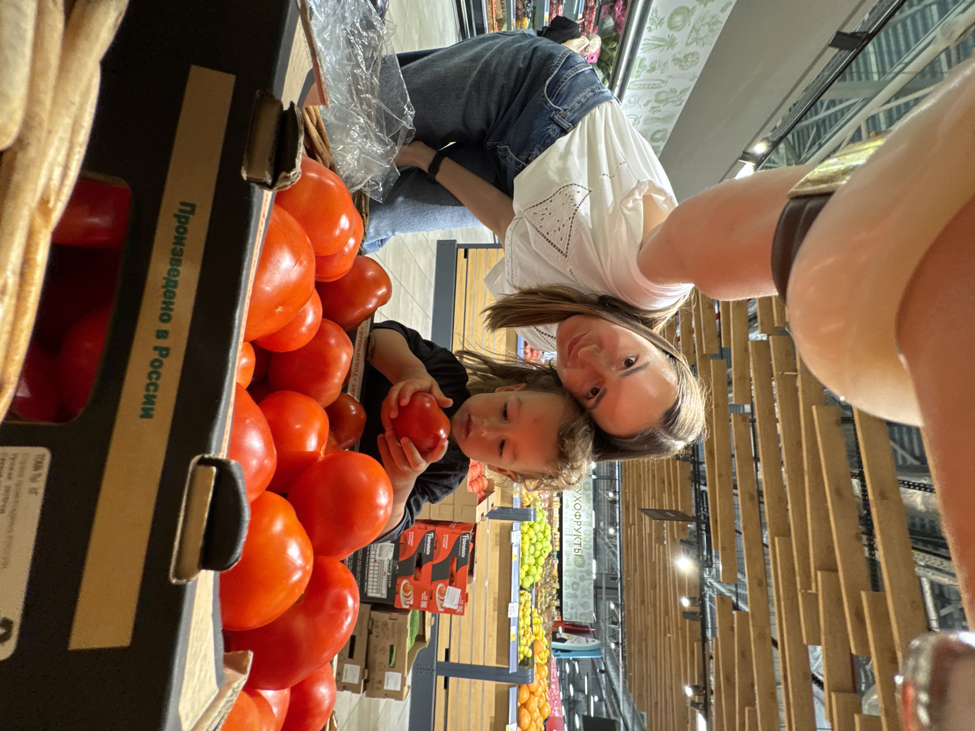 A woman and a young child are in a grocery store, looking at tomatoes. The woman is wearing a white t-shirt and blue jeans, and the child is wearing a black shirt. The child is holding a tomato. The grocery store has shelves of produce in the background, including fruits and vegetables. The location appears to be a supermarket with wooden shelving and fluorescent lighting, typical of many stores in Sochi, Russia. The image is a close-up shot, focusing on the woman, child, and a box of tomatoes in the foreground.