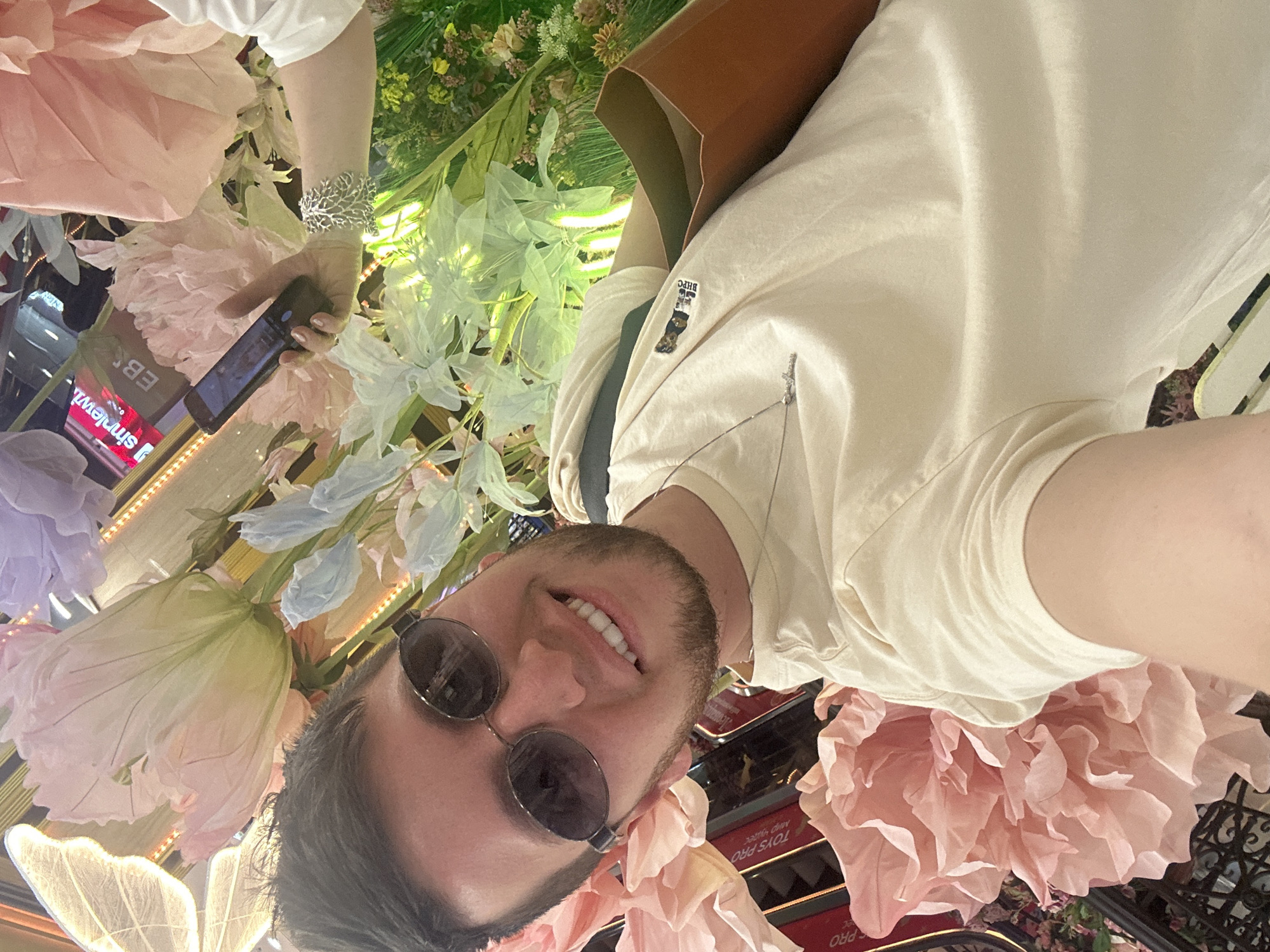 The image is a selfie taken by a man wearing sunglasses and a light-colored t-shirt, smiling at the camera. He has short dark hair and a beard. He is holding a brown shopping bag in his right hand. The background is decorated with large, artificial flowers in pastel colors, including pink, purple, and light blue. Behind the flowers, a sign with Russian text, "e simonei," is partially visible, along with a brightly lit display. The setting appears to be a retail or entertainment space, possibly a mall or a themed exhibition. Based on the text visible, the location is identified as Moscow, Russia.
