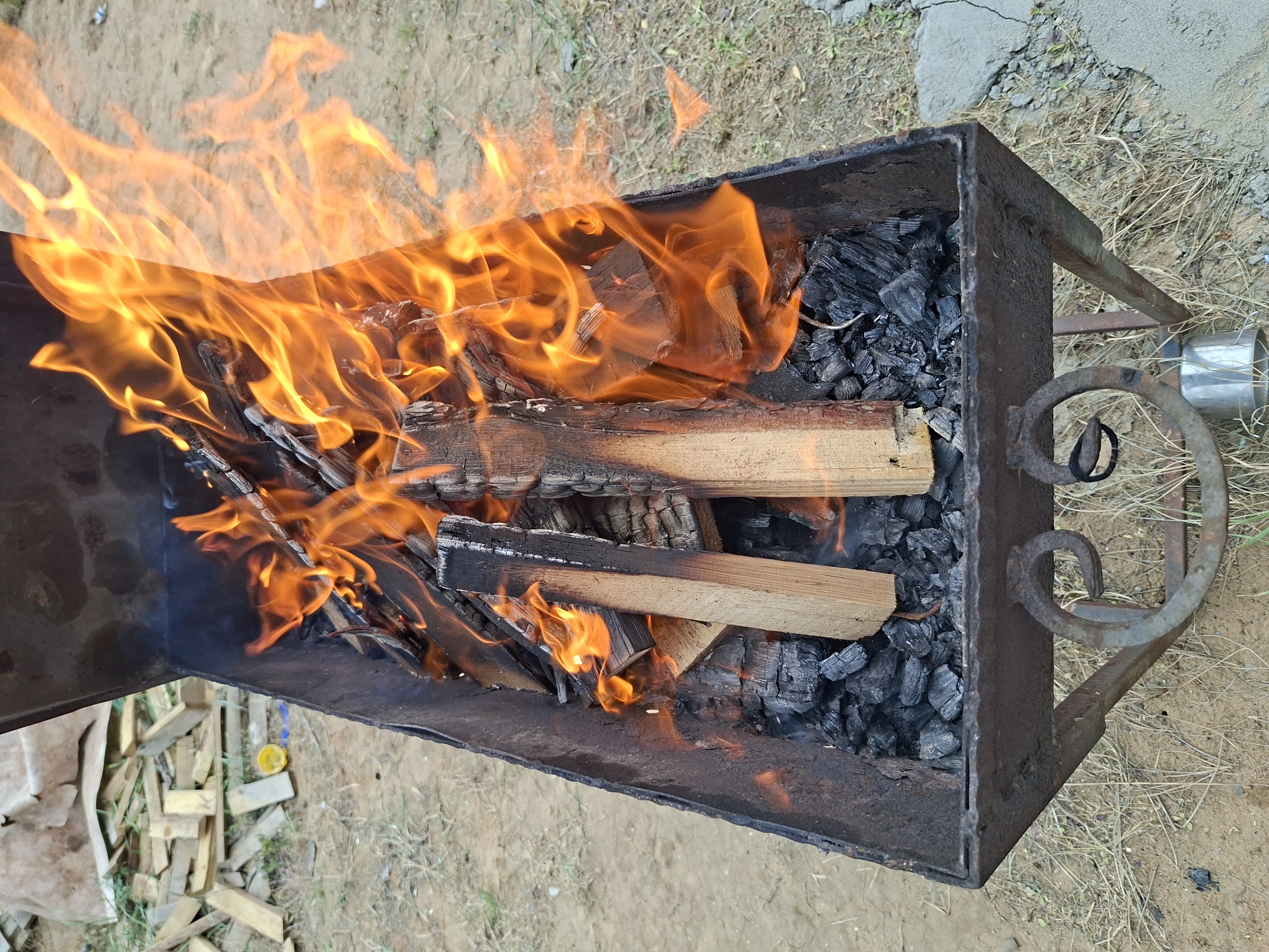 A top-down shot shows a rectangular brazier filled with burning wood and charcoal. The flames are bright orange and consume the wood. The charcoal beneath is dark and ashen. The brazier is rusty and sits on a metal frame with ornate, curved supports. The ground around the brazier is covered in dry grass and sandy soil. A silver cylindrical object is visible on the right side of the frame, near the brazier's supports.