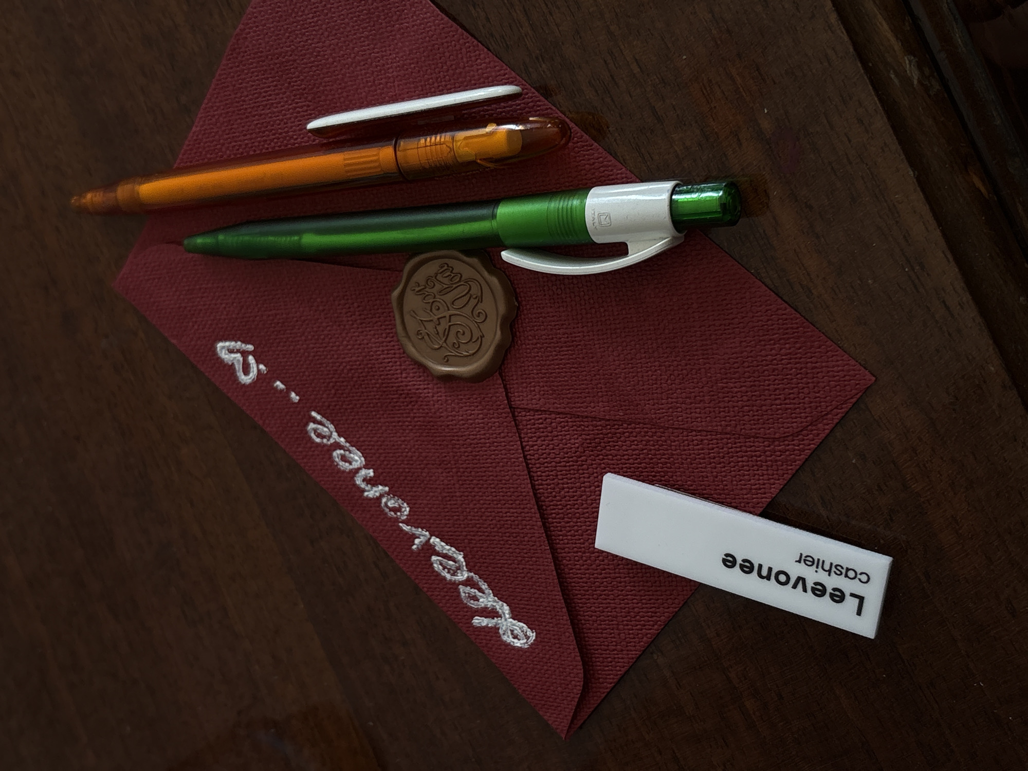 The media file captures a flat lay composition featuring stationery items arranged on a dark, rich brown wooden surface. The central object is a maroon-colored, textured paper, likely an envelope, positioned diagonally.
White embroidered text is visible along the left edge of the maroon paper, spelling "Leevonee", followed by a dashed line and the letter "B". A circular, brown wax seal with an ornate, possibly monogrammed, design is placed towards the top center of the paper.
Two pens rest on the maroon paper. The upper pen is a transparent orange ballpoint with a white cap. Below it is a green ballpoint pen featuring a silver-white clip. Closer inspection reveals faint text reading "ITAL" along with a logo on the silver part of the green pen.
In the bottom right corner, a white, rectangular tag is positioned. It displays the text "Leevonee" in a larger font and "cashier" in a smaller font, both oriented vertically.
The wooden surface shows clear grain patterns, suggesting a table or desk. The lighting is soft and even, indicating an indoor setting. No individuals are present, and the scene depicts a static arrangement of objects, possibly prepared for use or as an artistic display. While the provided context places this in Yerevan, Armenia, there are no specific visual cues within the image to confirm this location.