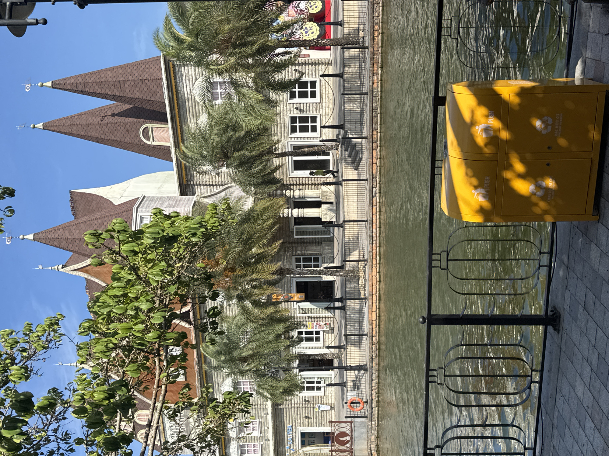 The image depicts a waterfront scene in Đà Nẵng, Vietnam. A building with distinctive architecture, featuring pointed roofs and light-colored wooden cladding, stands along the water's edge. Palm trees line the building, and a metal railing separates the walkway from the water. A yellow recycling bin is positioned on the walkway near the railing. The water appears calm and reflects the surrounding structures. The sky is clear and blue.